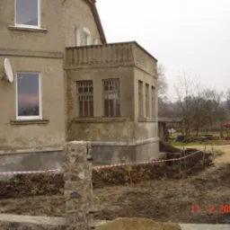 Przygotowanie terenu wokół starego domu z elewacją w kolorze piaskowym, widoczne wykopy zabezpieczone taśmą ostrzegawczą, sugerujące prace ziemne przy fundamentach.