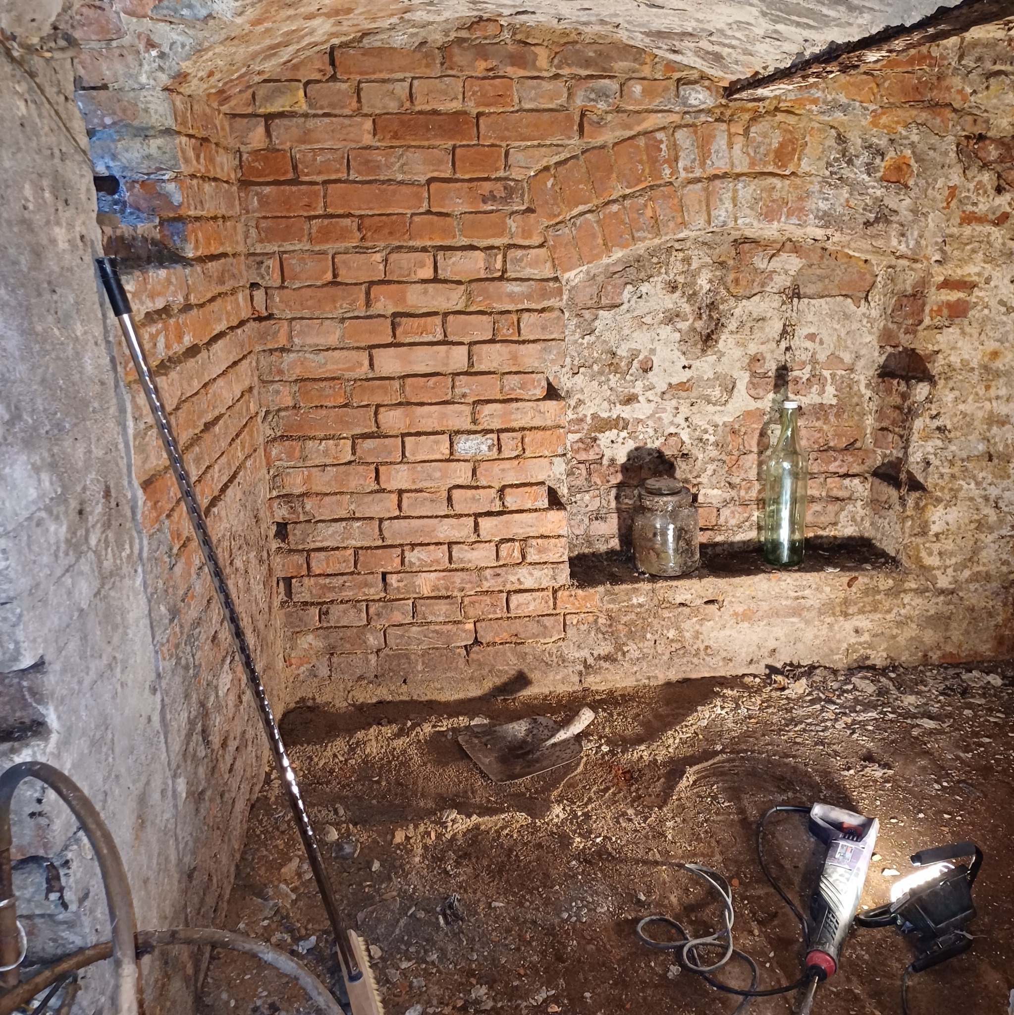 Piwnica z ceglanymi ścianami, widoczne ślady wilgoci, narzędzia budowlane i oświetlenie, sugerujące prace osuszające lub izolacyjne.