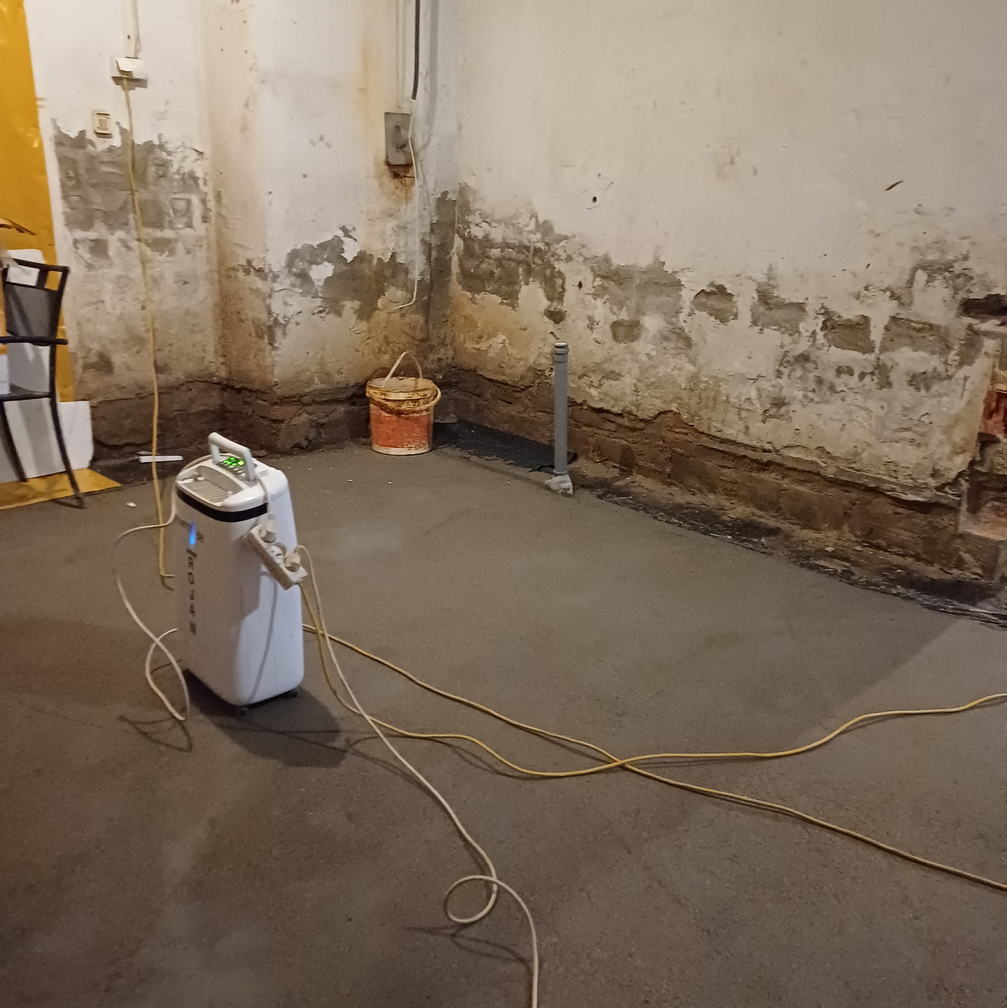 Biały osuszacz powietrza stoi na świeżo wylanym betonie w piwnicy z wilgotnymi ścianami, widoczny kabel zasilający i wiadro w tle.
