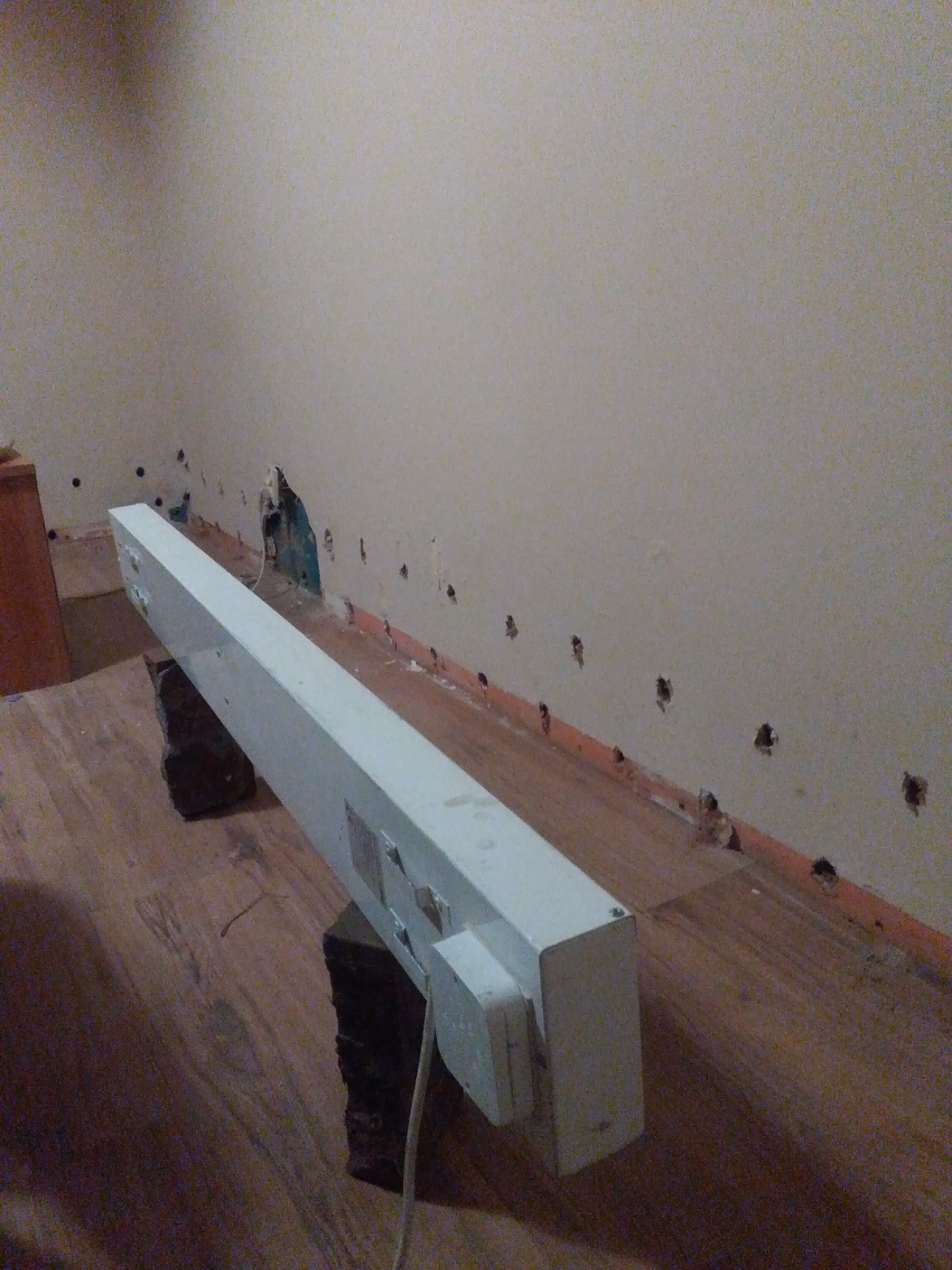 Ściana z nawierconymi otworami wzdłuż podłogi, z widocznymi śladami wilgoci i uszkodzeniami tynku, oświetlona tymczasową lampą LED umieszczoną na kamieniach.