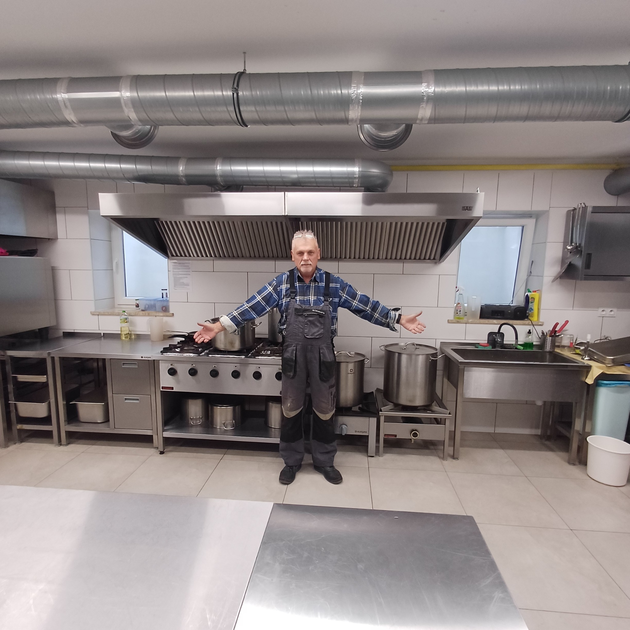 Wnętrze kuchni przemysłowej po remoncie z nową wentylacją. Mężczyzna w kombinezonie prezentuje efekt prac. Widoczne stalowe meble i garnki.