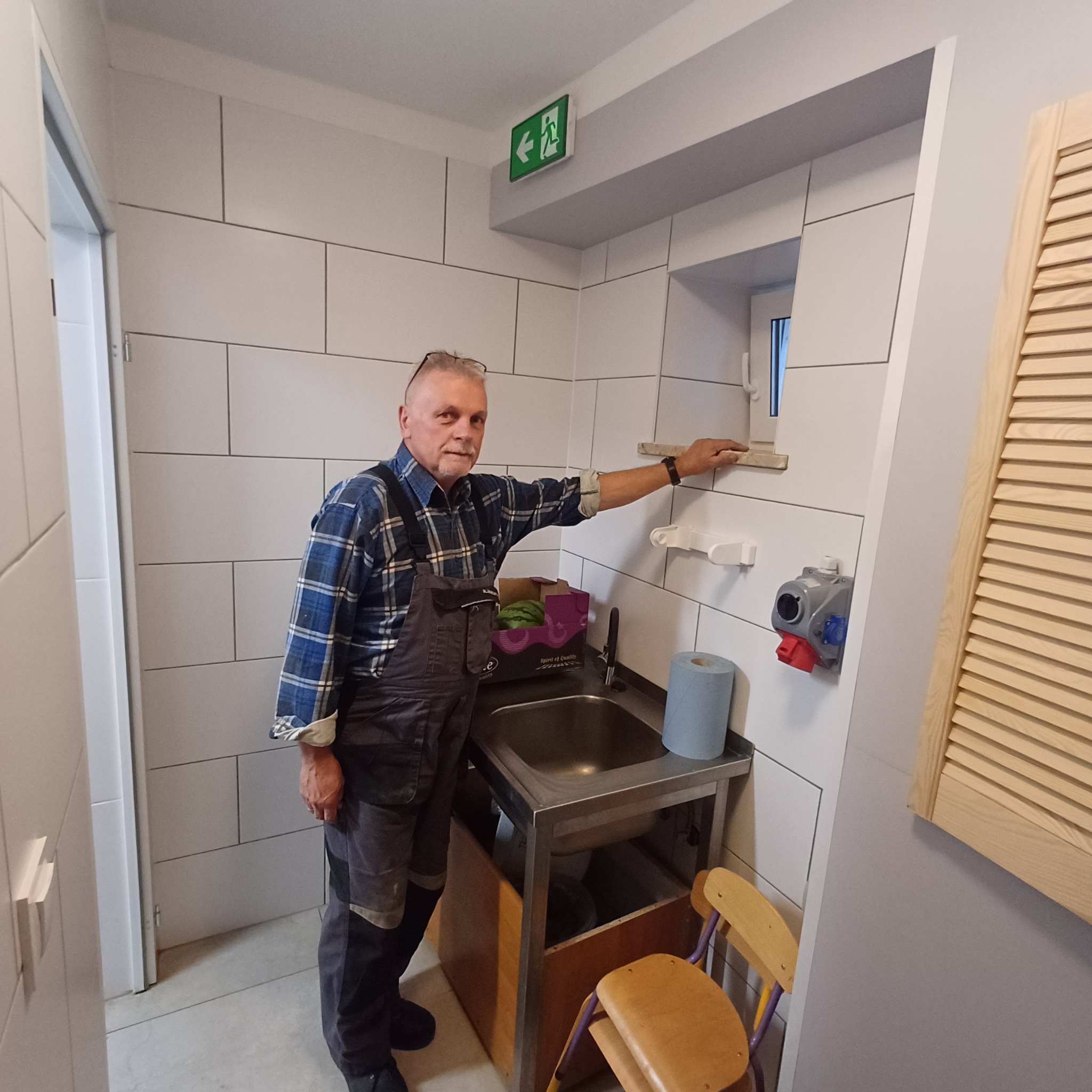 Pracownik w kombinezonie prezentuje efekt osuszania ściany w pomieszczeniu sanitarnym w Zielonej Górze. Widoczna umywalka i świeżo położone białe płytki.