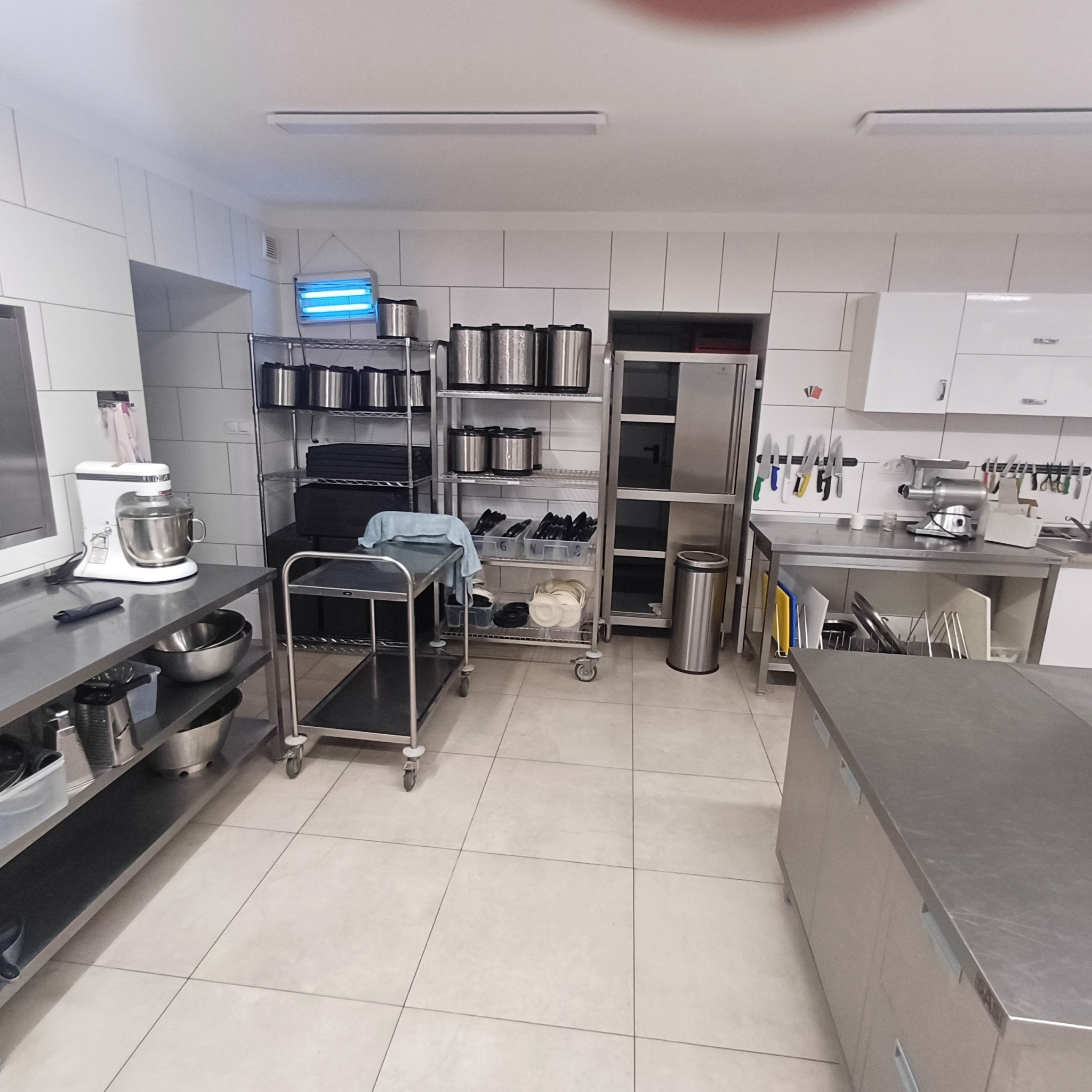Profesjonalna kuchnia gastronomiczna: stal nierdzewna, sprzęt, czyste płytki. Widoczny mikser, krajalnica, regały i wózek transportowy.