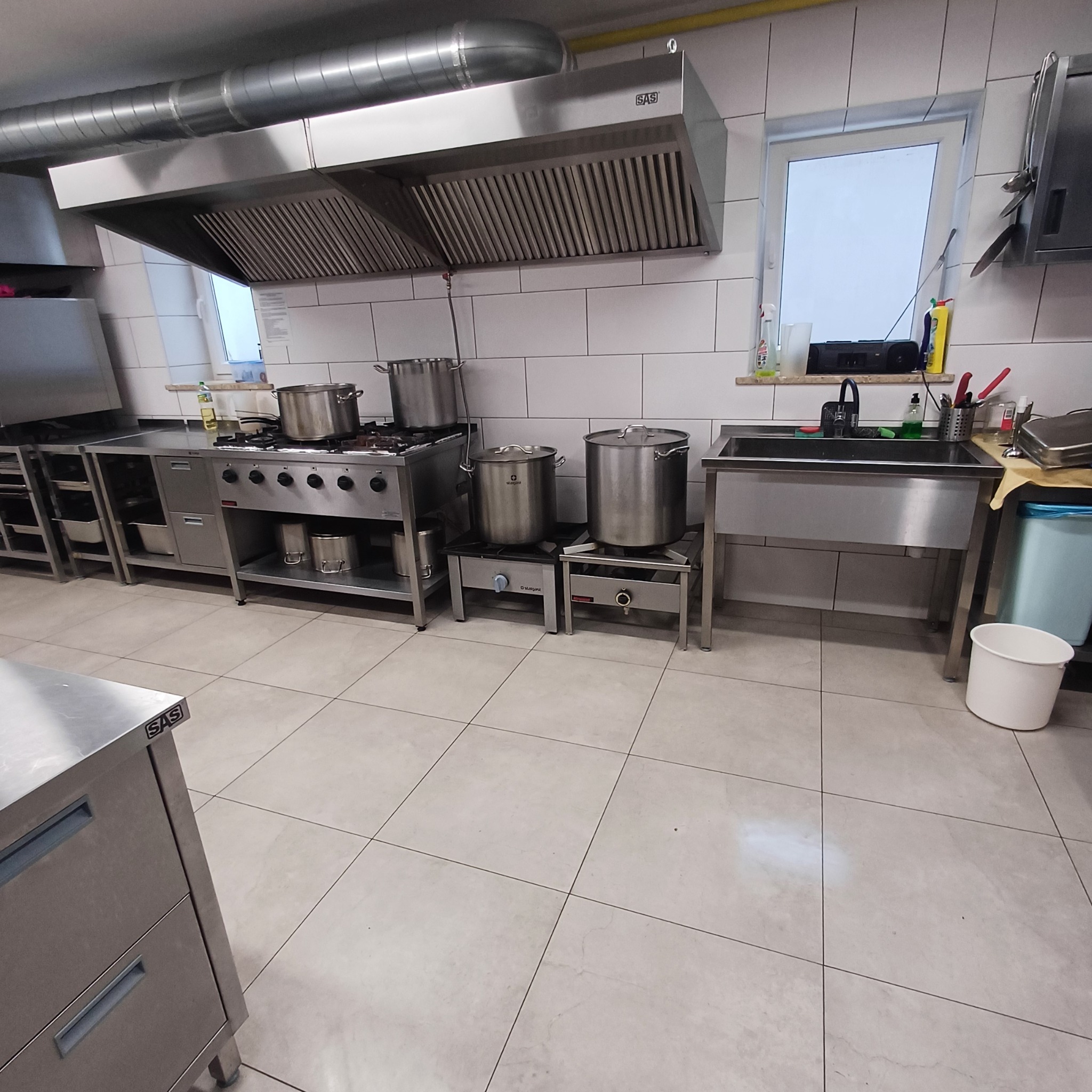 Profesjonalna kuchnia gastronomiczna ze stali nierdzewnej: okap, kuchenka, garnki, zlew. Białe płytki na ścianach i podłodze. Widok na okno.