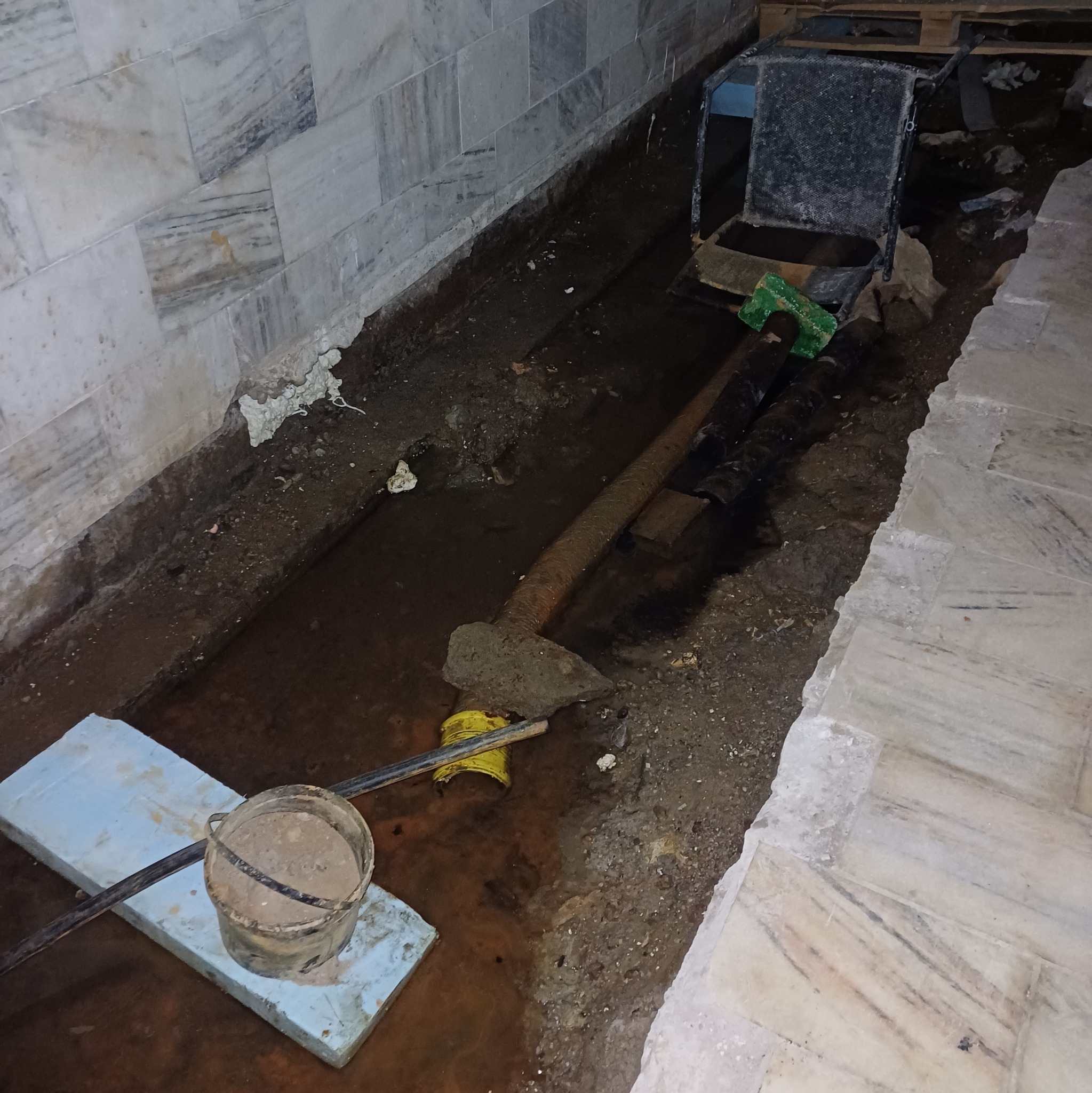 Wykopany rów wewnątrz budynku z widocznymi elementami instalacji i narzędziami, prawdopodobnie w trakcie prac związanych z naprawą lub modernizacją.