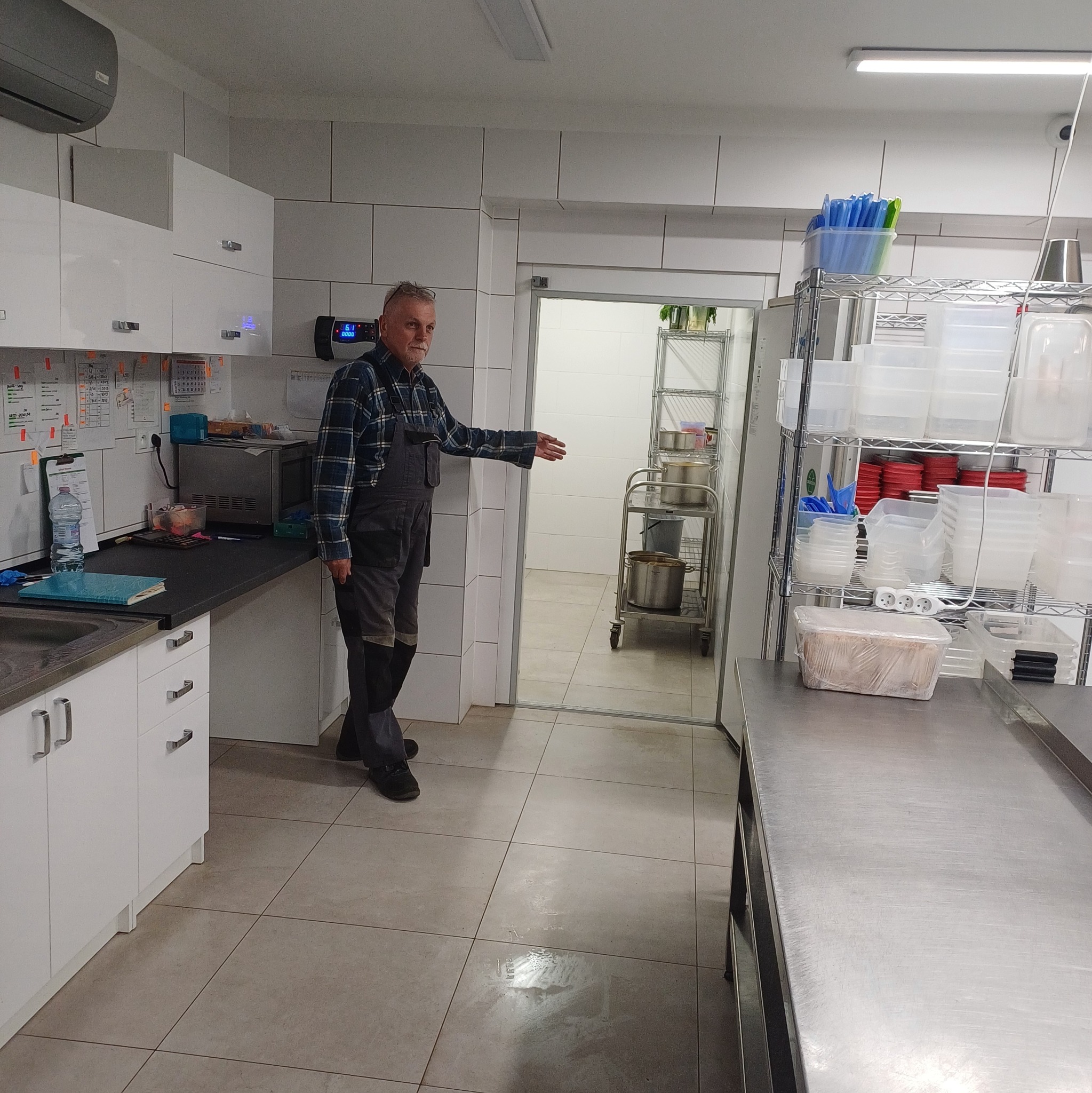 Pracownik w kuchni przemysłowej wskazuje na osuszone pomieszczenie. Widoczne białe płytki, stalowe regały i sprzęty kuchenne. Czyste, jasne wnętrze.