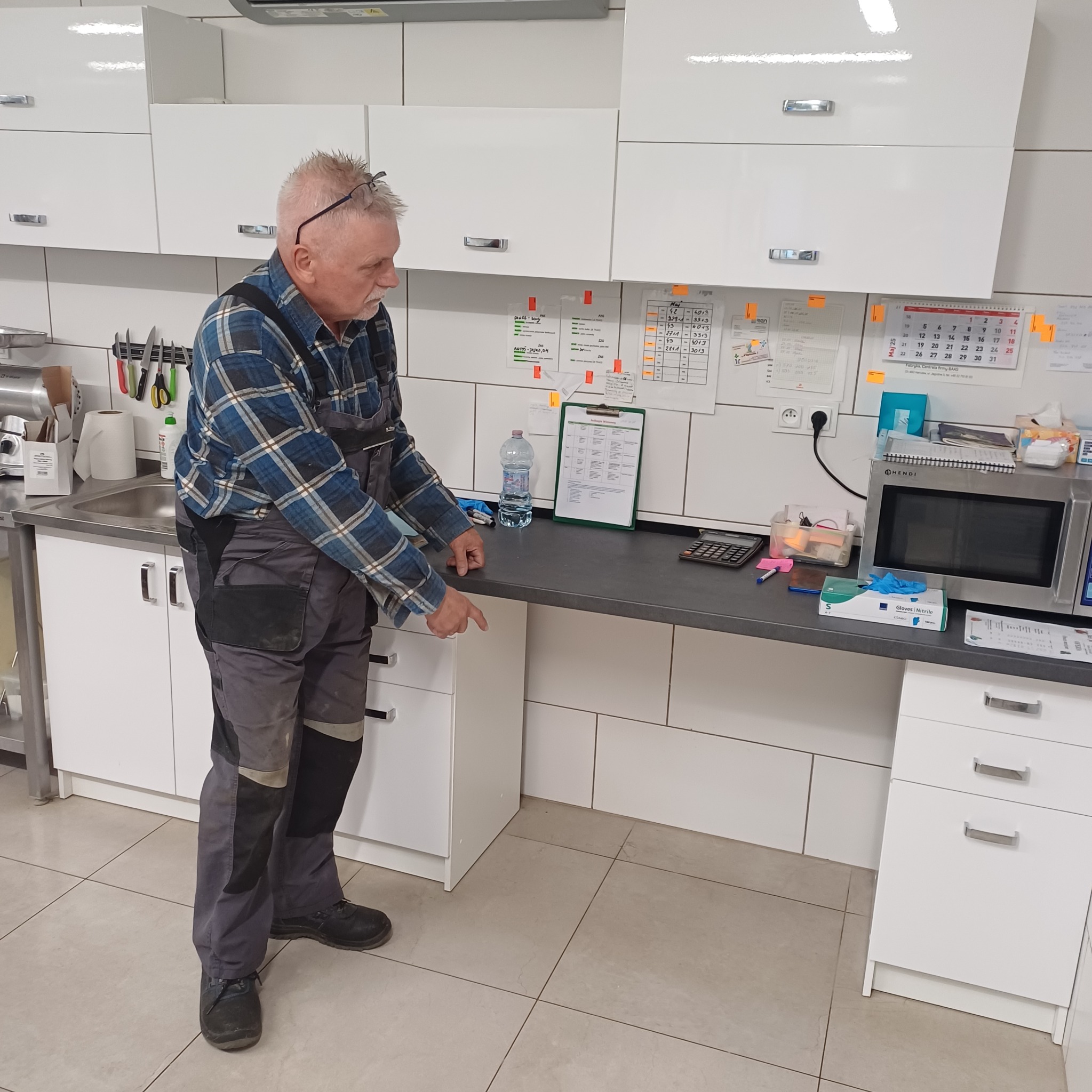 Mężczyzna w roboczym ubraniu, w pomieszczeniu z białymi szafkami, wskazuje na blat. Na blacie kalkulator, rękawice ochronne i inne przedmioty.