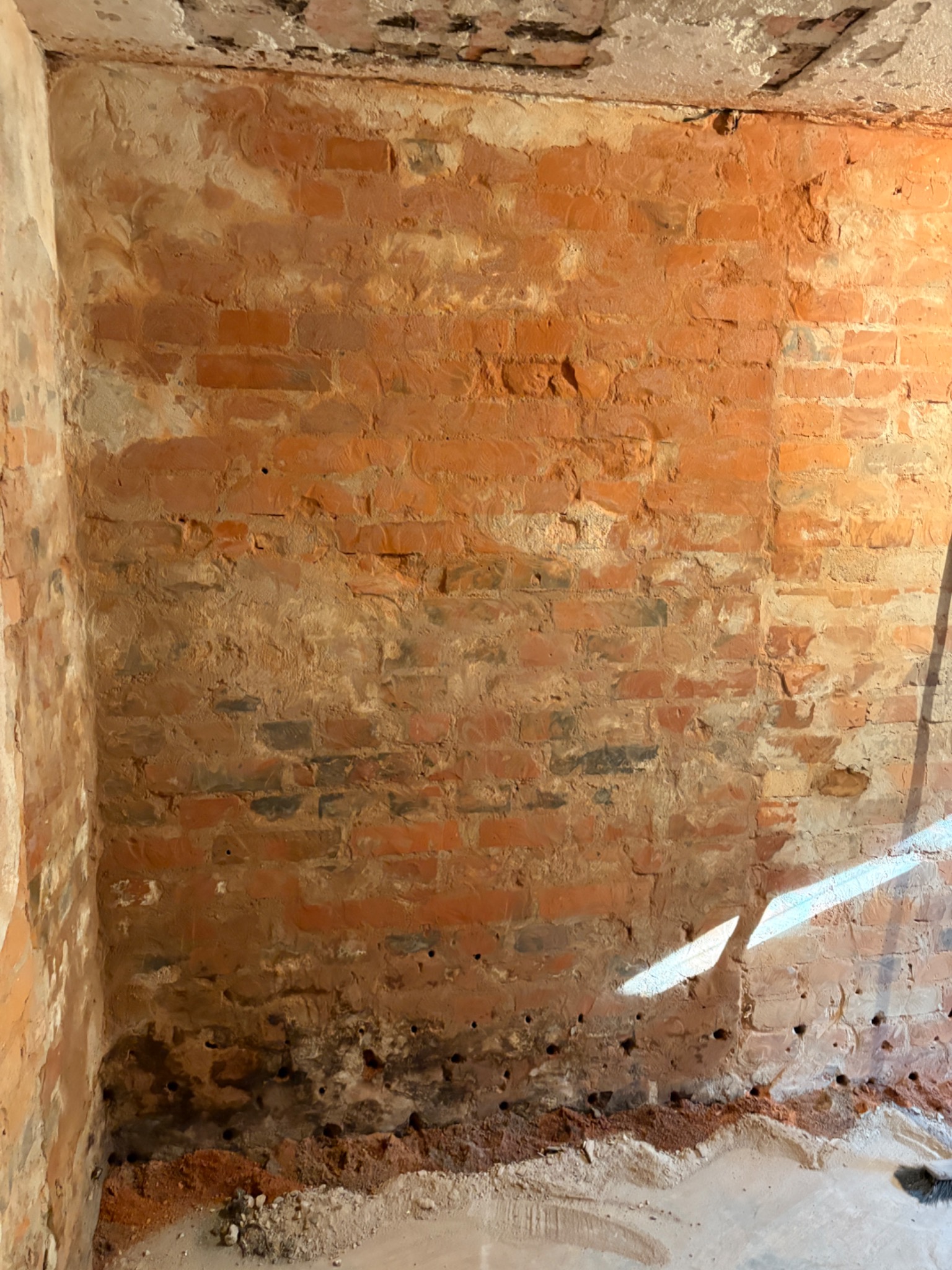 Ceglana ściana z widocznymi śladami wilgoci i wykwitami, wskazująca na problem z izolacją i potrzebę osuszania. Widoczne otwory w dolnej części ściany.