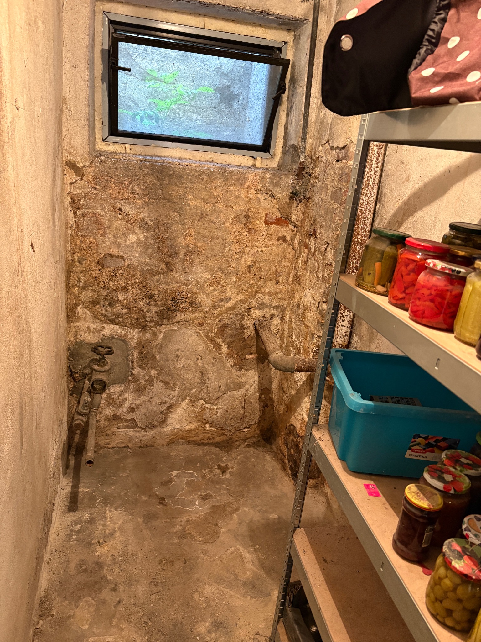 Wilgotna piwnica z oknem, ścianą z odpadającym tynkiem i rurami. Na półce słoiki z przetworami. Widoczne ślady zawilgocenia i potencjalnej potrzeby osuszania.