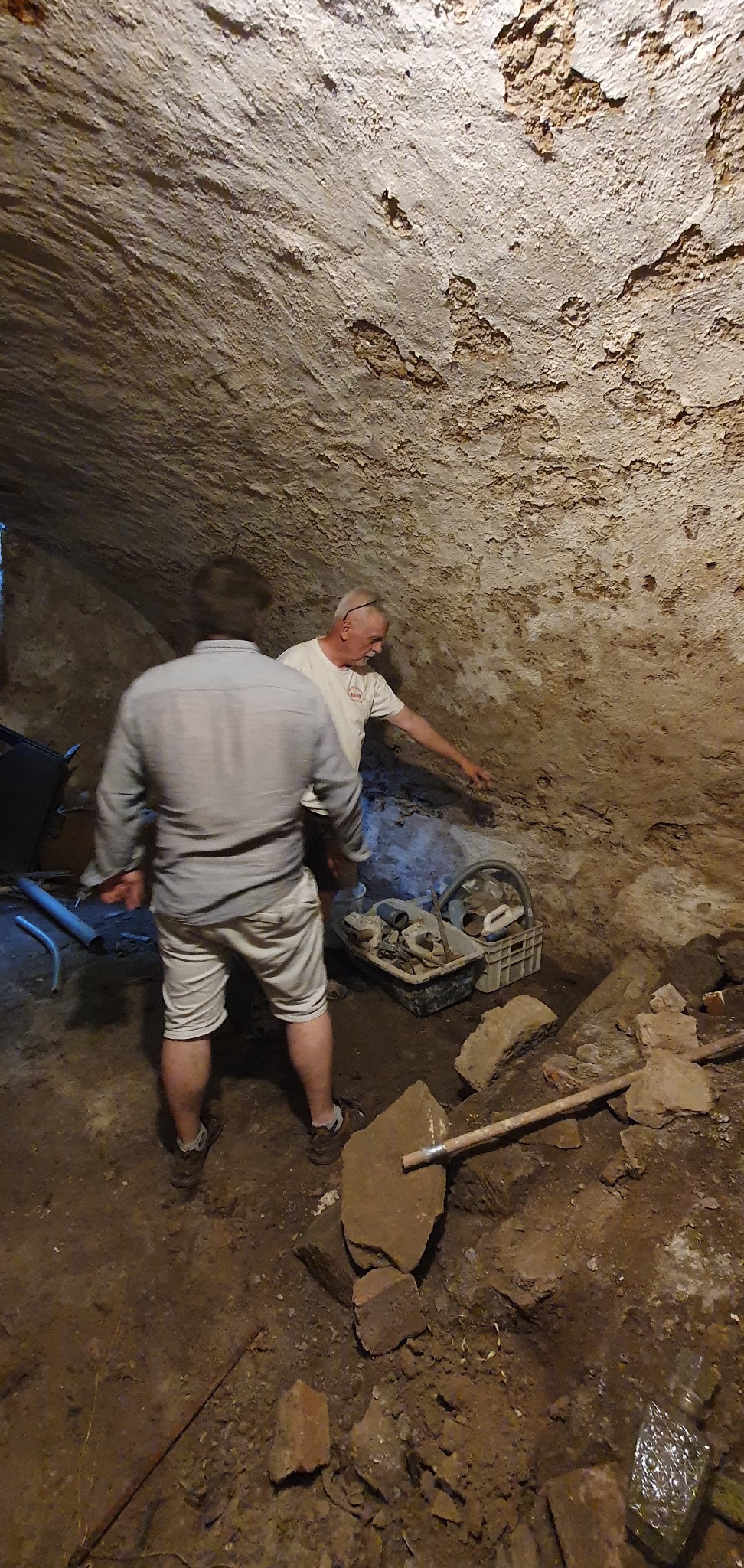 Inspekcja zawilgoconej piwnicy z narzędziami i dwoma mężczyznami, z których jeden wskazuje na ścianę z widocznymi śladami wilgoci.