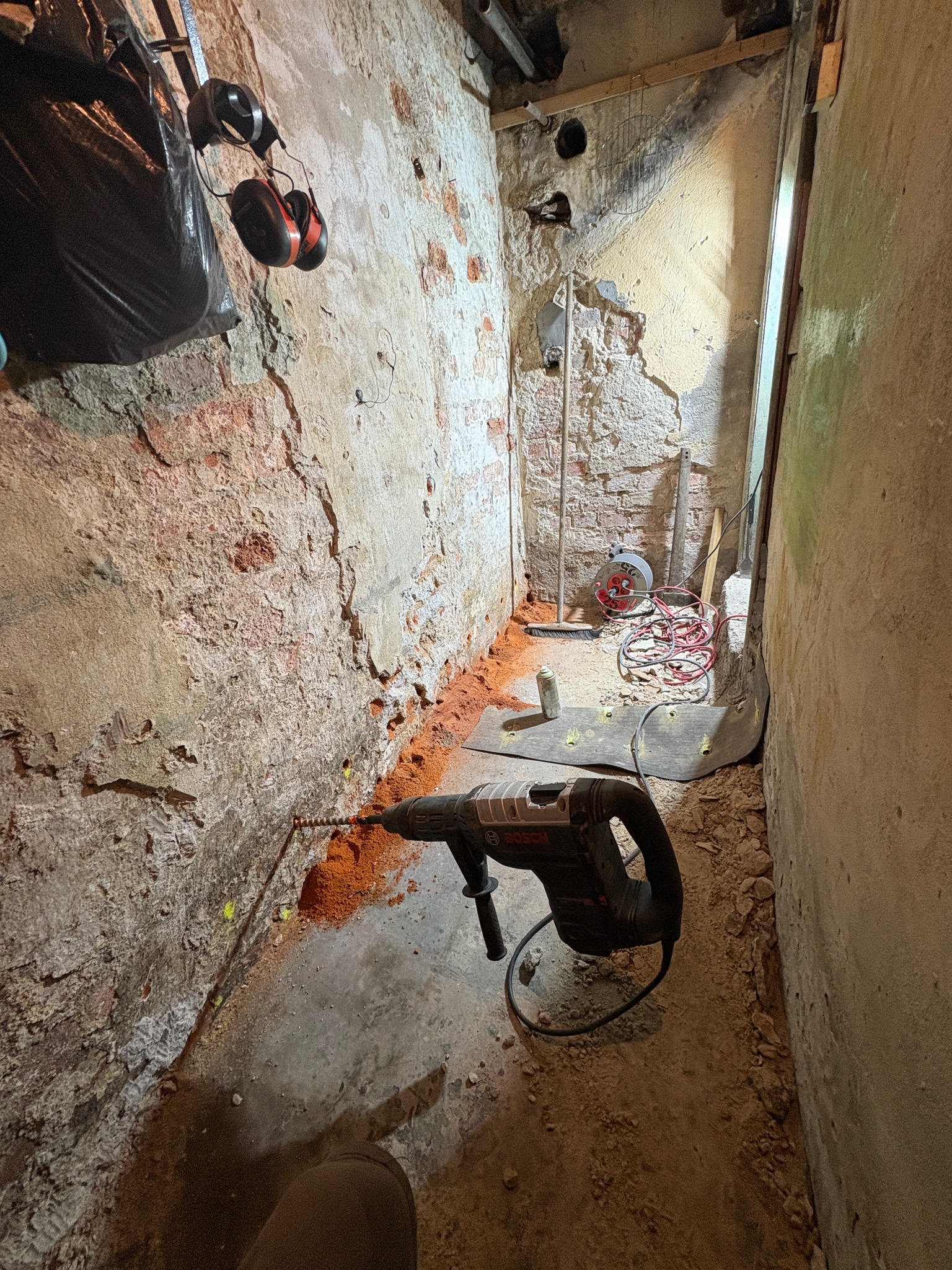 Wykonywanie otworów w ścianie przy podłodze w pomieszczeniu z odsłoniętą cegłą, widoczny młot udarowy Bosch i pył po wierceniu. Prace remontowe w wąskim pomieszczeniu.
