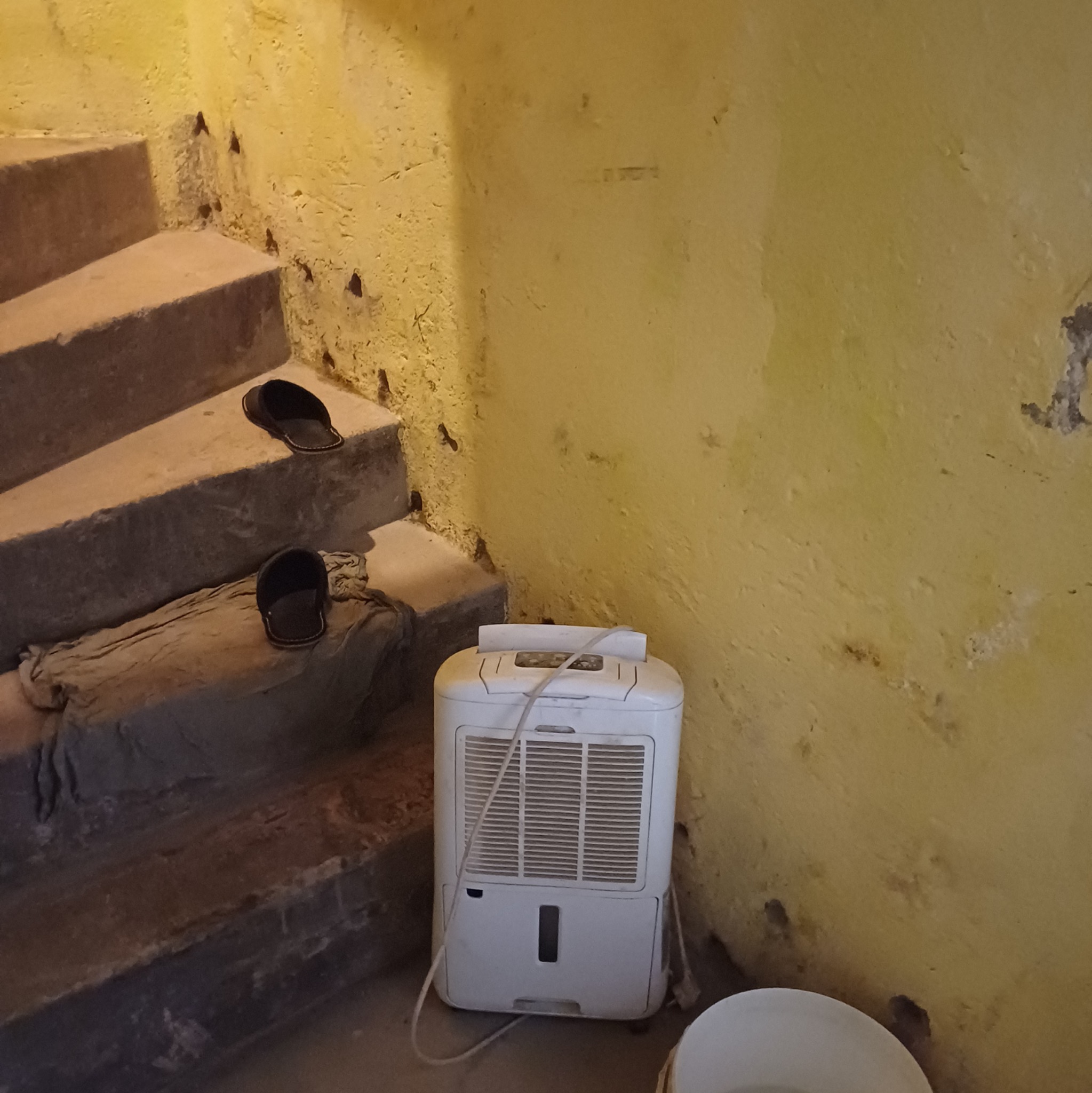 Biały osuszacz powietrza stoi przy żółtej ścianie i betonowych schodach z parą czarnych klapek. Widoczne ślady wilgoci na ścianie.