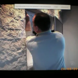 Mężczyzna w trakcie montażu dekoracyjnej ściany z kamienia imitującego piaskowiec wewnątrz pomieszczenia, widoczne fragmenty ramy okiennej i telewizora.