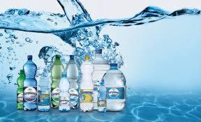 Różne rodzaje i wielkości butelek wody mineralnej z etykietami producenta, ustawione na tle błękitnej wody z efektem rozprysku.