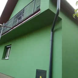 Świeżo pomalowana, jasnozielona elewacja domu z ciemnoszarą rynną i balkonem z metalową balustradą. Widoczna skrzynka gazowa na ścianie i fragment rusztowania.