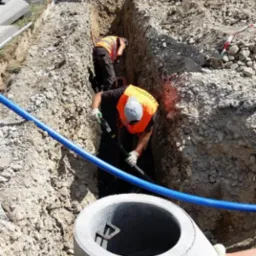 Montaż betonowego kręgu studni przez dwóch pracowników w wykopie, z widocznym mini-koparką i rurami. Praca w toku, teren budowy.