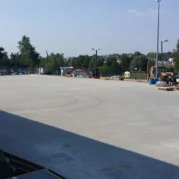 Rozległy, betonowy plac z częściowo ułożoną kostką brukową w tle, widoczny sprzęt budowlany i pracownicy. Jasne światło słoneczne, drzewa i budynki w tle.