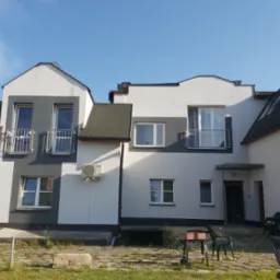 Dwukondygnacyjny budynek z ocieploną elewacją w kolorach bieli i szarości, z balkonami i klimatyzatorami na ścianach, widoczny z poziomu trawnika.