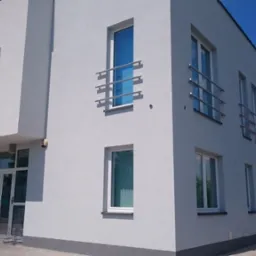 Nowoczesny, dwukondygnacyjny budynek biurowy z szarą elewacją, metalowymi balustradami przy oknach i przeszklonym wejściem. W tle widoczny fragment sąsiedniego budynku z czerwoną dachówką...