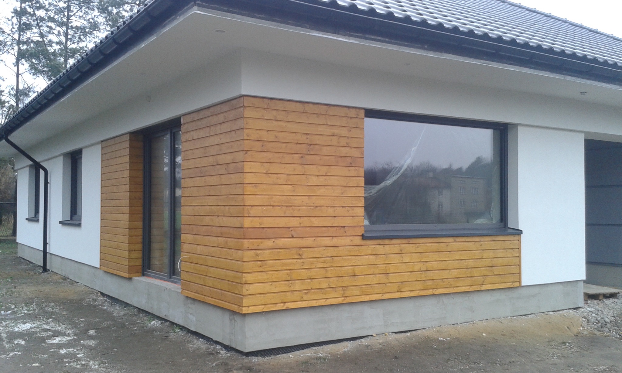 Nowoczesny dom jednorodzinny z fragmentem elewacji wykończonym drewnianymi panelami w ciepłym odcieniu, widoczne okno z czarną ramą i fragment dachu pokrytego szarą dachówką.