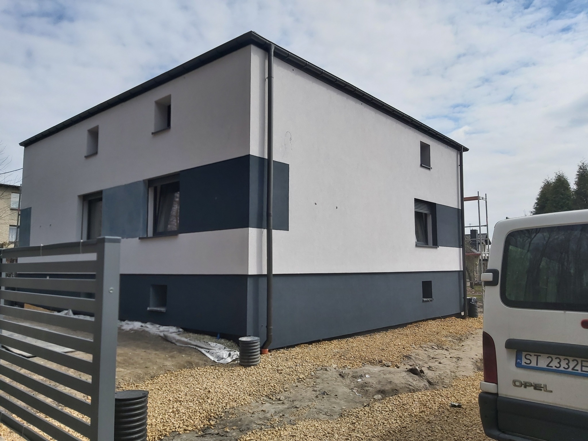 Nowoczesny dom jednorodzinny po remoncie elewacji, z dwukolorową fasadą: biała górna część i grafitowa dolna, widoczne rynny i obróbki blacharskie, teren wokół domu wysypany kamieniem.