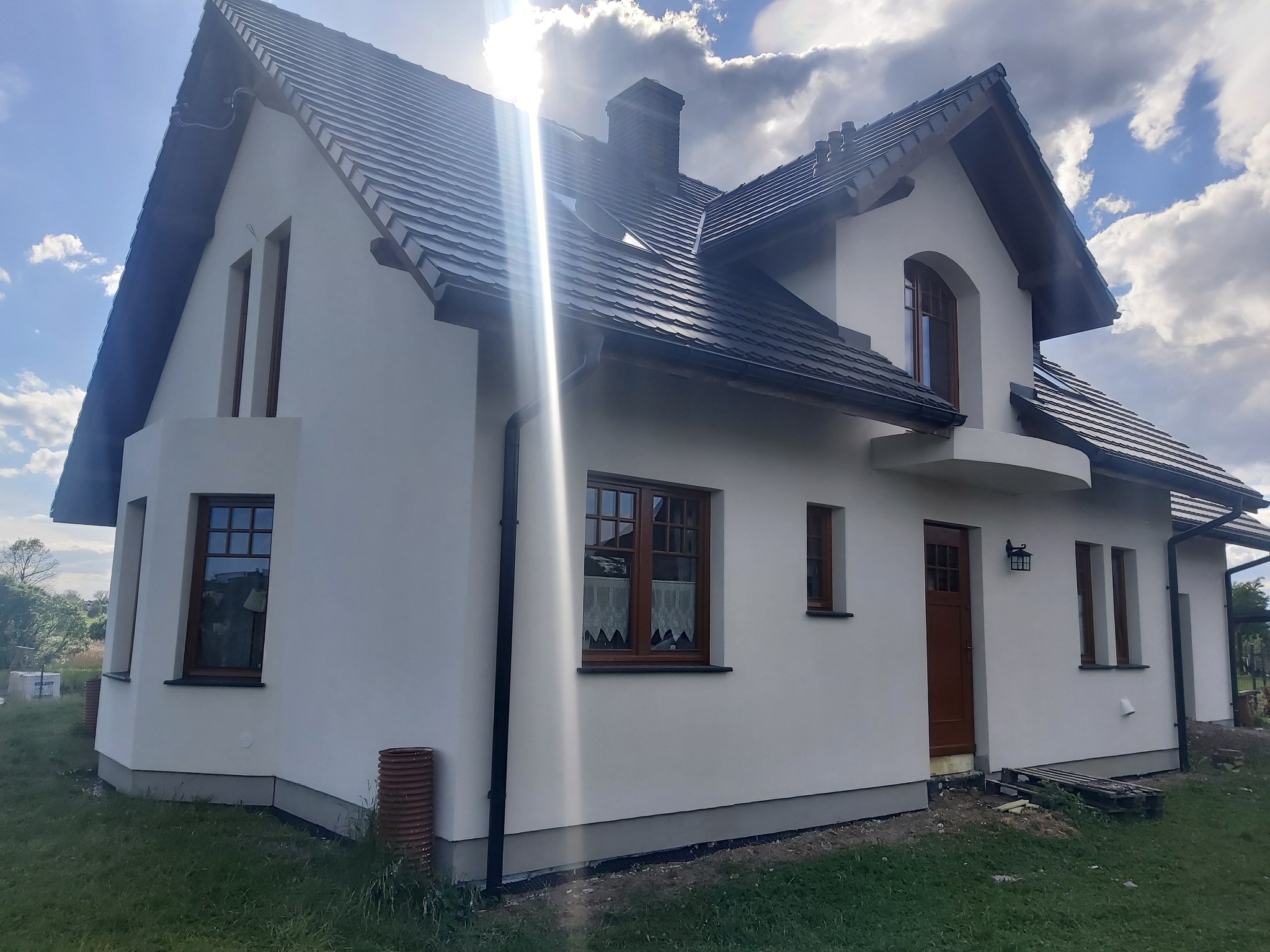 Nowo wykończony dom jednorodzinny z białą elewacją, brązowymi oknami i ciemną dachówką, uchwycony w słoneczny dzień z widocznymi detalami wykończenia.