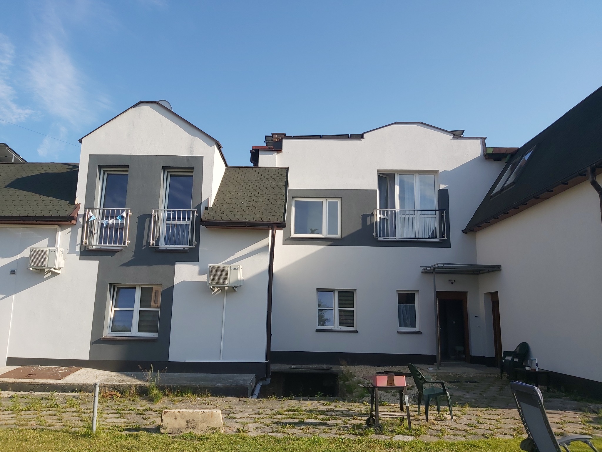 Dwukondygnacyjny budynek z ocieploną elewacją w kolorach bieli i szarości, z balkonami i klimatyzatorami na ścianach, widoczny z poziomu trawnika.