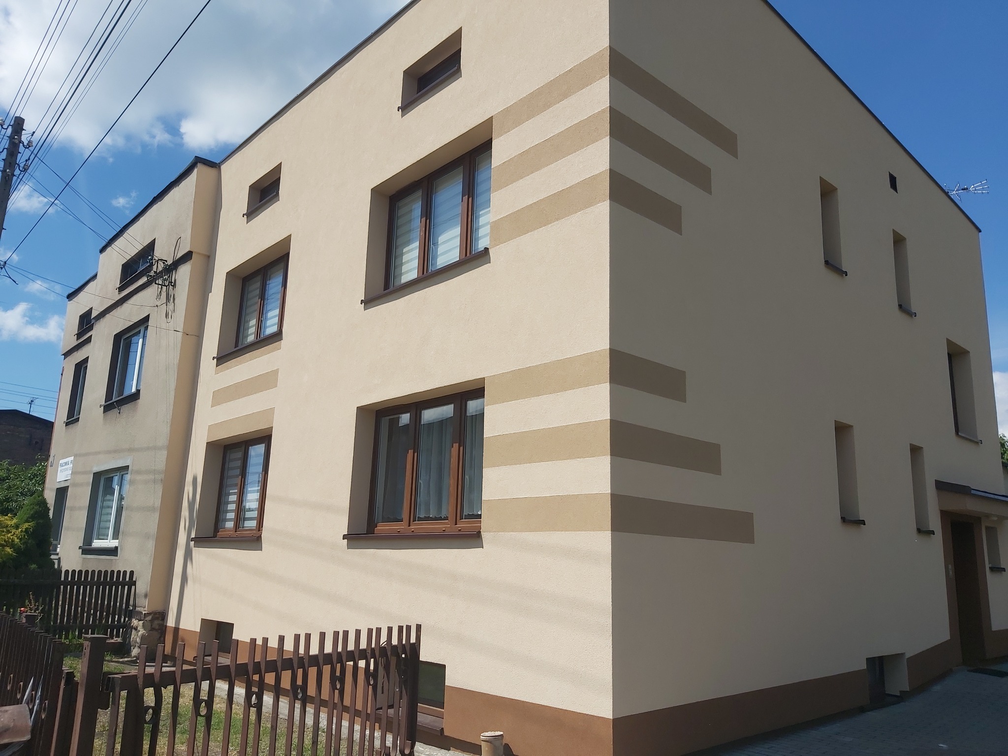 Dwukondygnacyjny budynek z beżową elewacją ozdobioną poziomymi pasami w odcieniach brązu, widok z zewnątrz w słoneczny dzień.