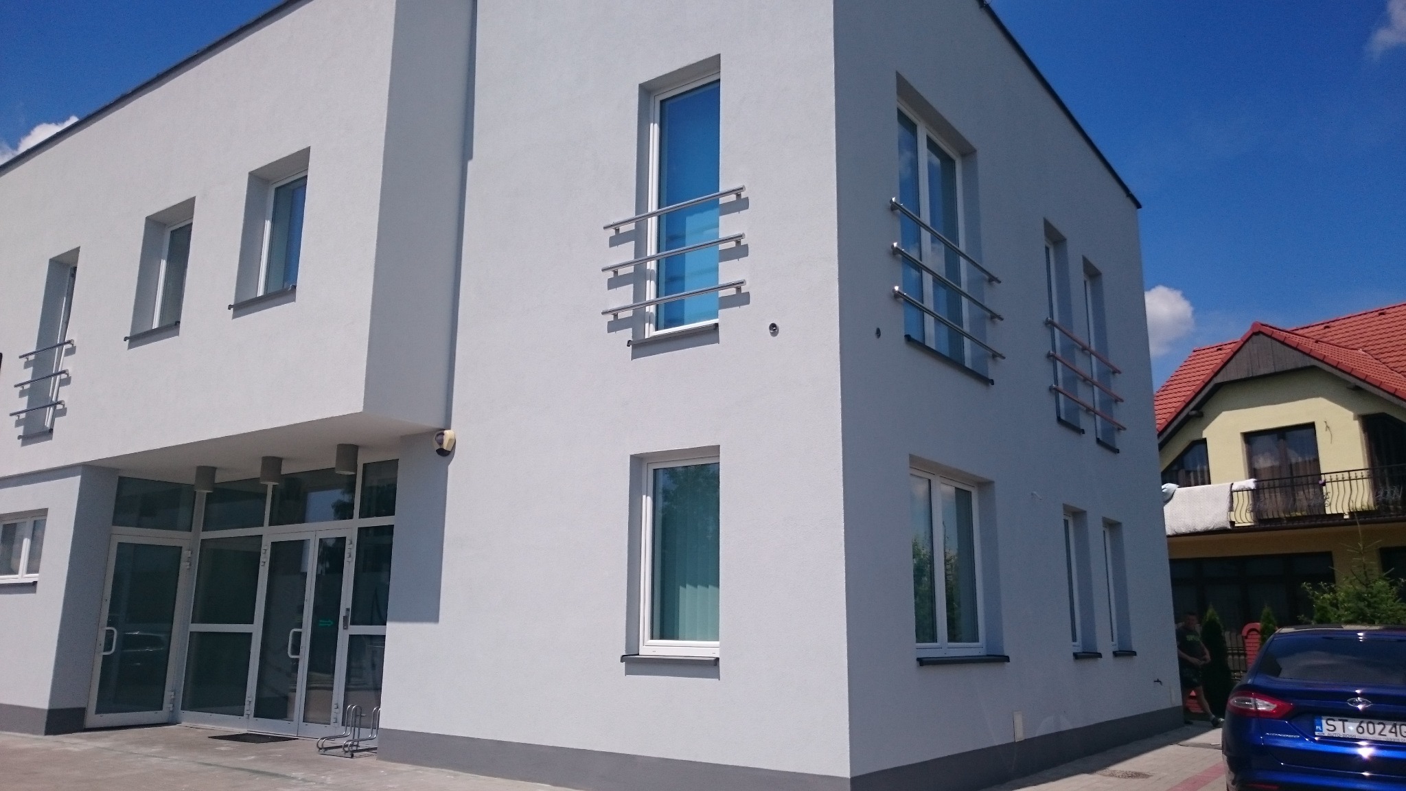 Nowoczesny, dwukondygnacyjny budynek biurowy z szarą elewacją, metalowymi balustradami przy oknach i przeszklonym wejściem. W tle widoczny fragment sąsiedniego budynku z czerwoną dachówką...