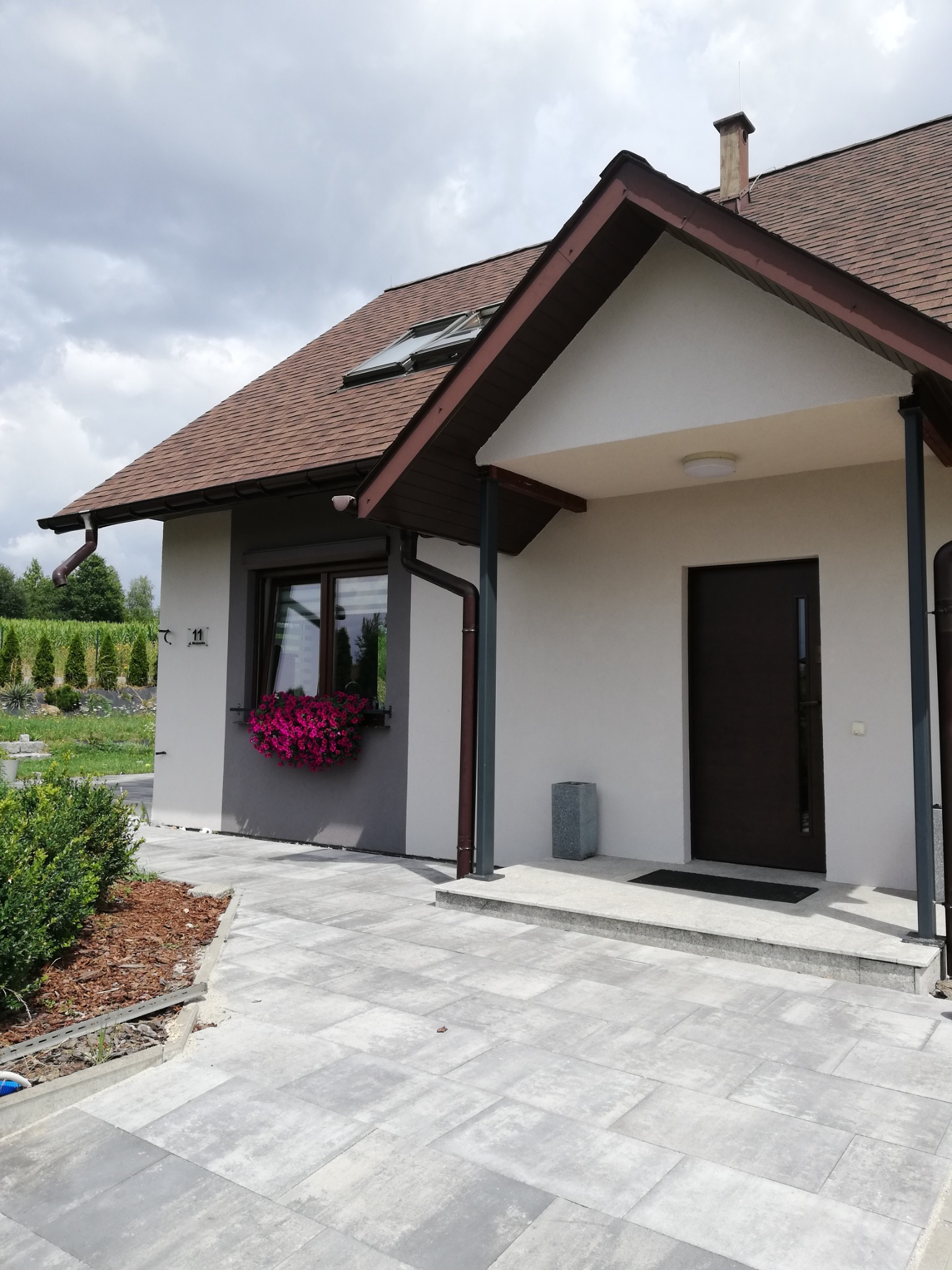 Ukończony dom jednorodzinny z brązowym dachem, szarą elewacją w dolnej części i jasną w górnej, z rynnami w kolorze dachu i skrzynką z kwiatami pod oknem.