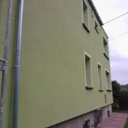 Elewacja budynku po tynkowaniu na zielono, z widocznymi oknami w jasnych obramowaniach i rurą spustową. Widoczna ścieżka z czerwonej kostki brukowej.