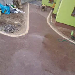 Wykończona, brązowa nawierzchnia z kostki brukowej z jasną obwódką, prowadząca do budynku. Widoczne przygotowane materiały i narzędzia.