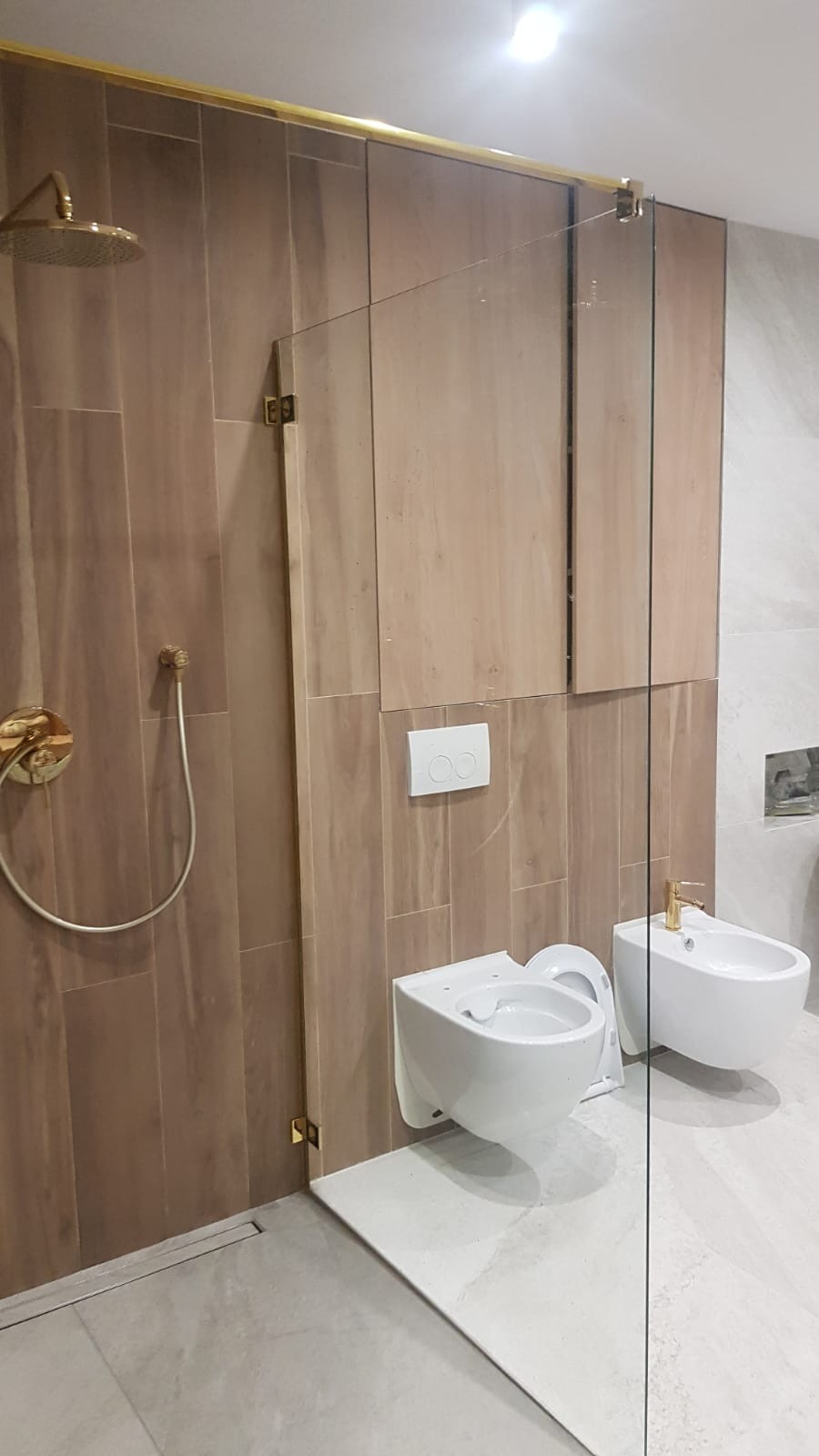 Nowoczesna łazienka z drewnopodobnymi płytkami, złotą armaturą i szklaną kabiną prysznicową typu walk-in, prezentująca minimalistyczny design i wysoką jakość wykończenia.