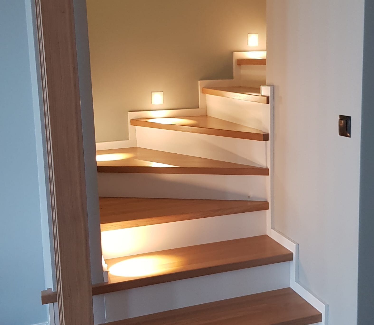 Drewniane schody z białymi podstopnicami i oświetleniem punktowym w ścianie, widoczne z dołu, z fragmentem ościeżnicy po lewej stronie.