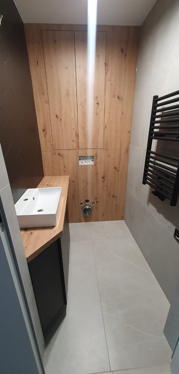 Nowoczesna łazienka po remoncie: drewniana zabudowa ściany z ukrytymi drzwiczkami, biała umywalka na drewnianym blacie, szary grzejnik drabinkowy i płytki w odcieniach szarości.