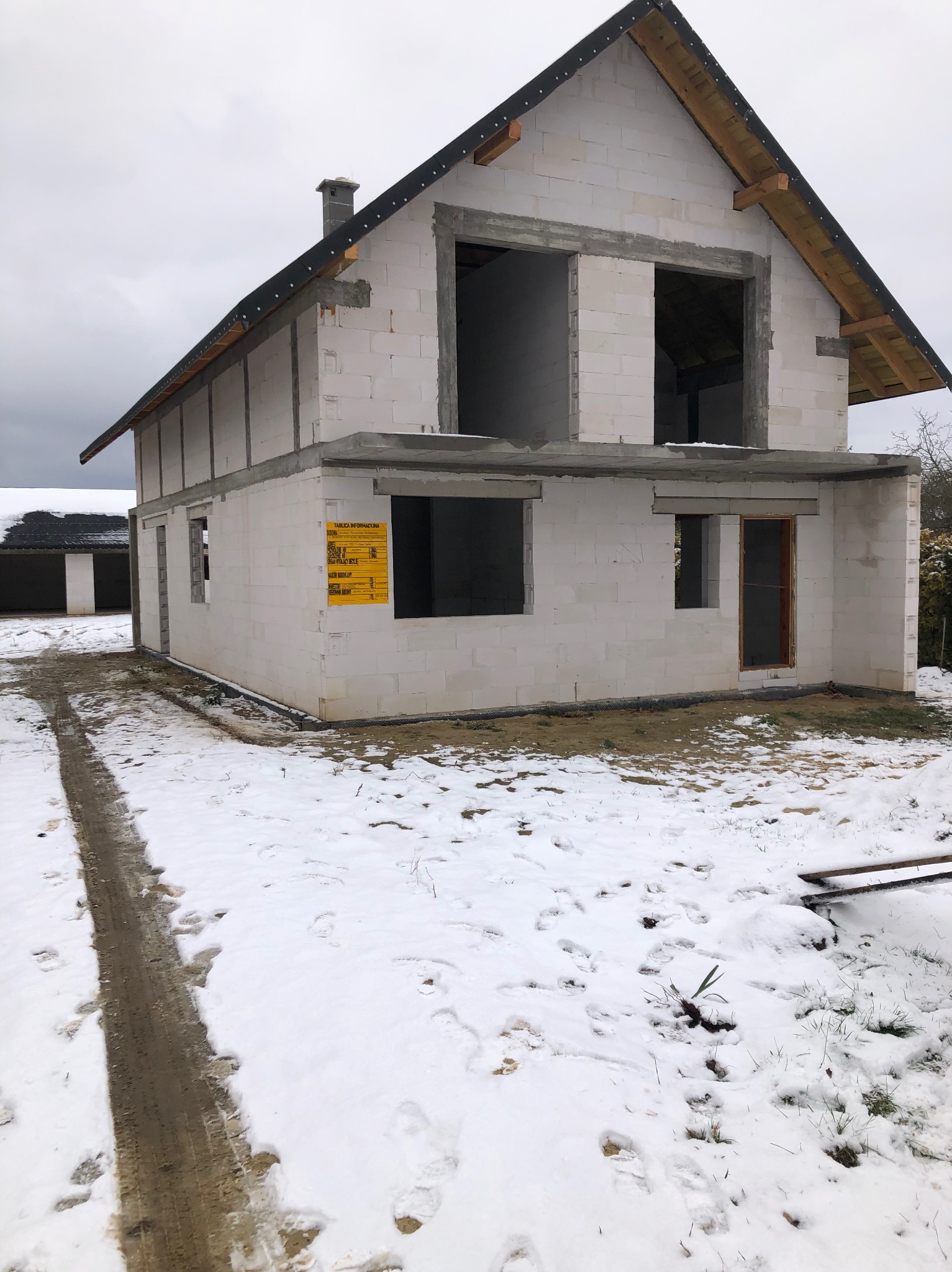Surowy stan domu murowanego z betonu komórkowego w zimowej scenerii, widoczna tablica informacyjna budowy, ślady opon na śniegu.