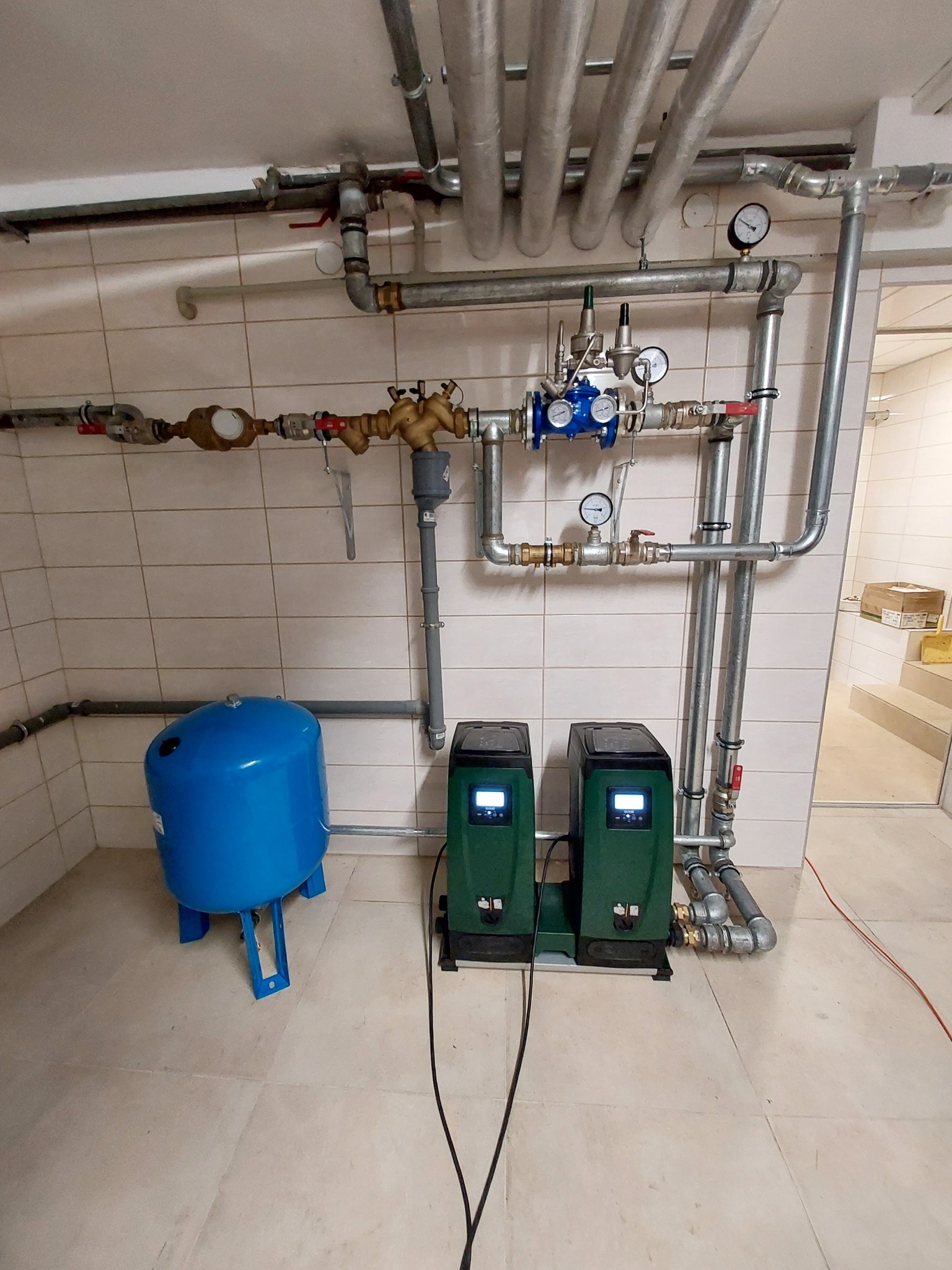 Zestaw hydroforowy z dwoma pompami obok niebieskiego zbiornika ciśnieniowego, widoczne rury instalacji wodnej z zaworami i manometrami, na ścianie płytki ceramiczne.