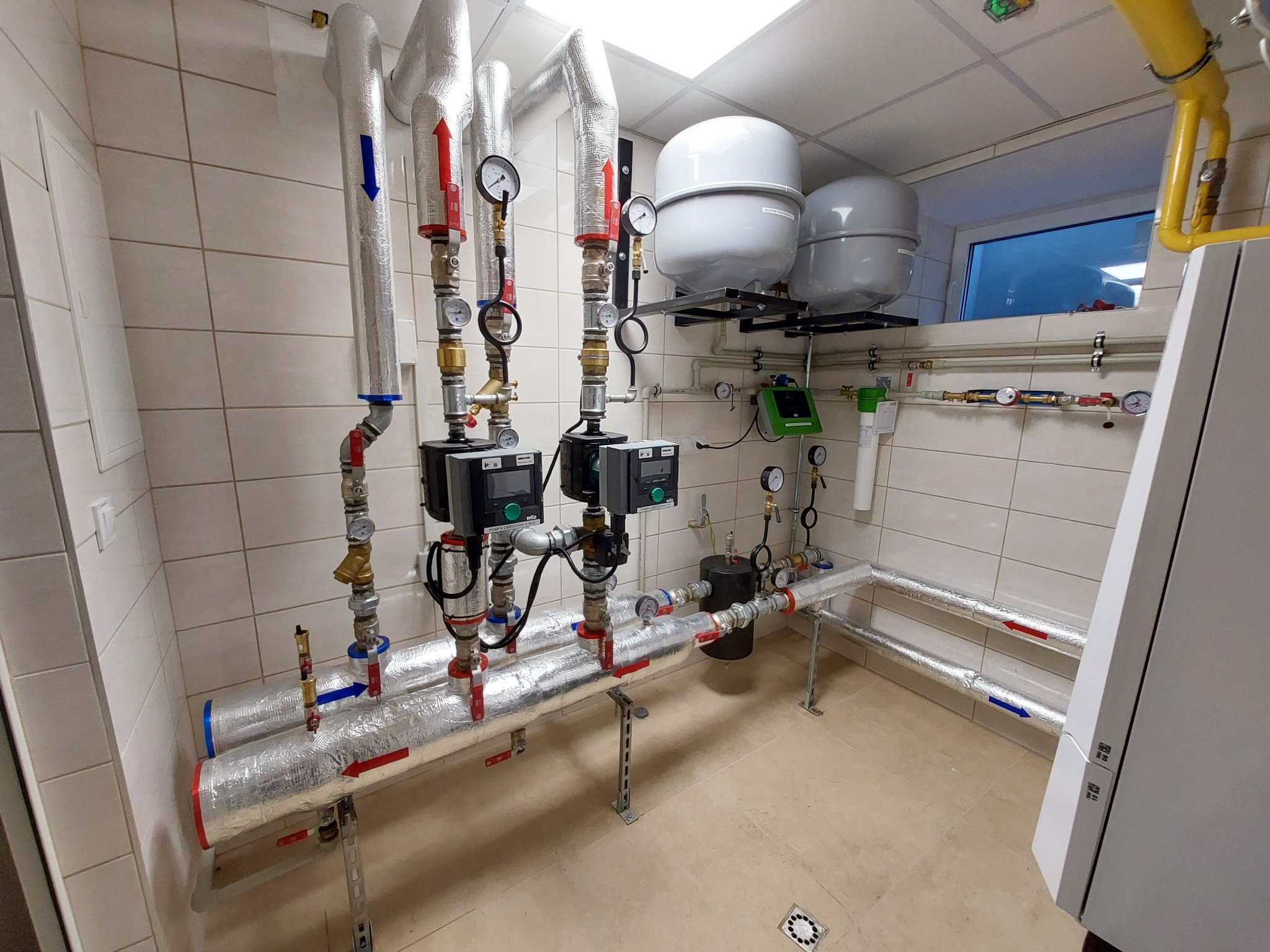 Węzeł cieplny w kotłowni z rurami izolowanymi folią aluminiową, manometrami, zaworami, pompami obiegowymi Wilo i zbiornikami wyrównawczymi na tle białych płytek ceramicznych.