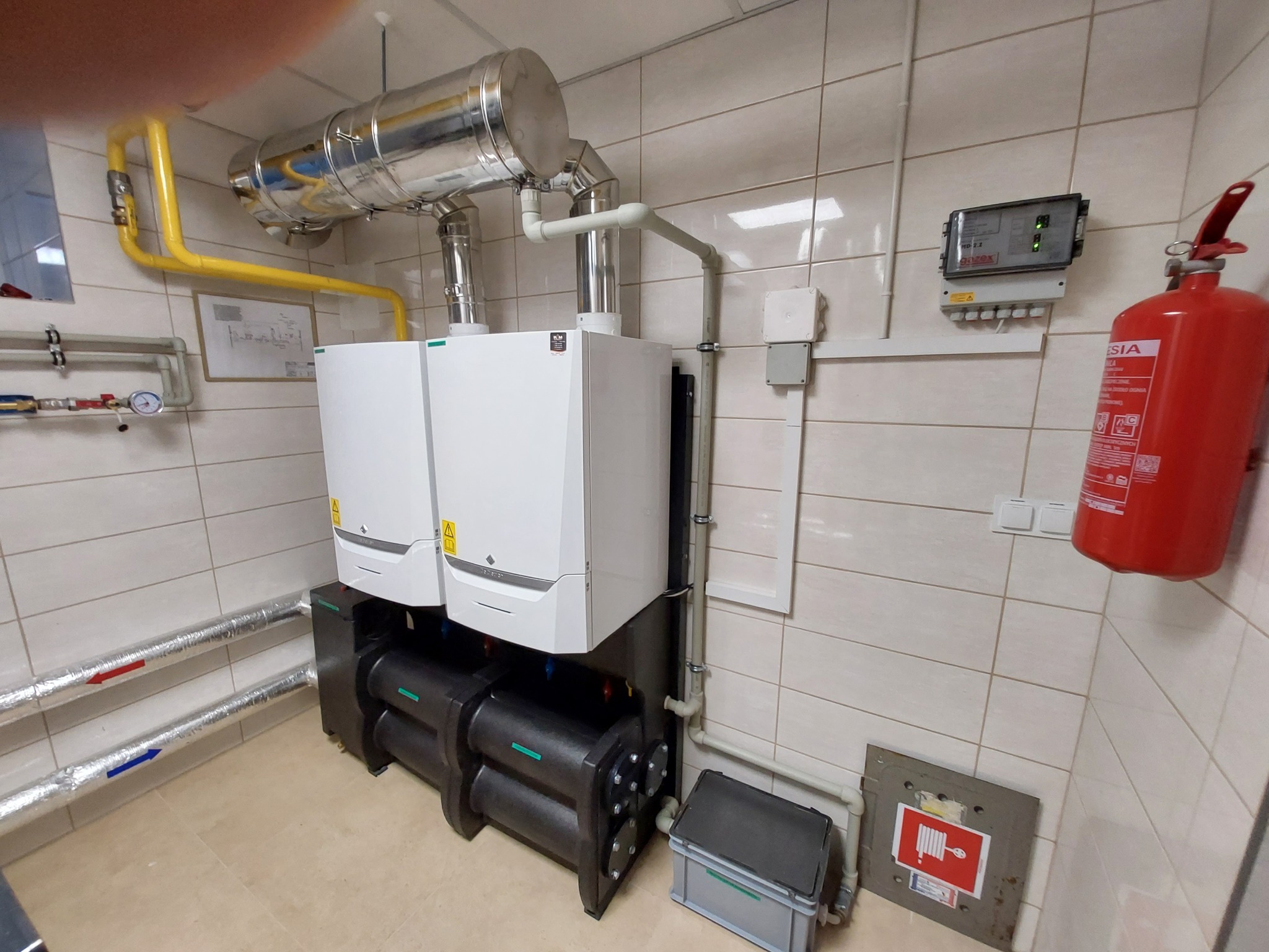 Dwa białe kotły grzewcze zamontowane na czarnej podstawie w pomieszczeniu z kafelkami, widoczne rury, sterownik i gaśnica.