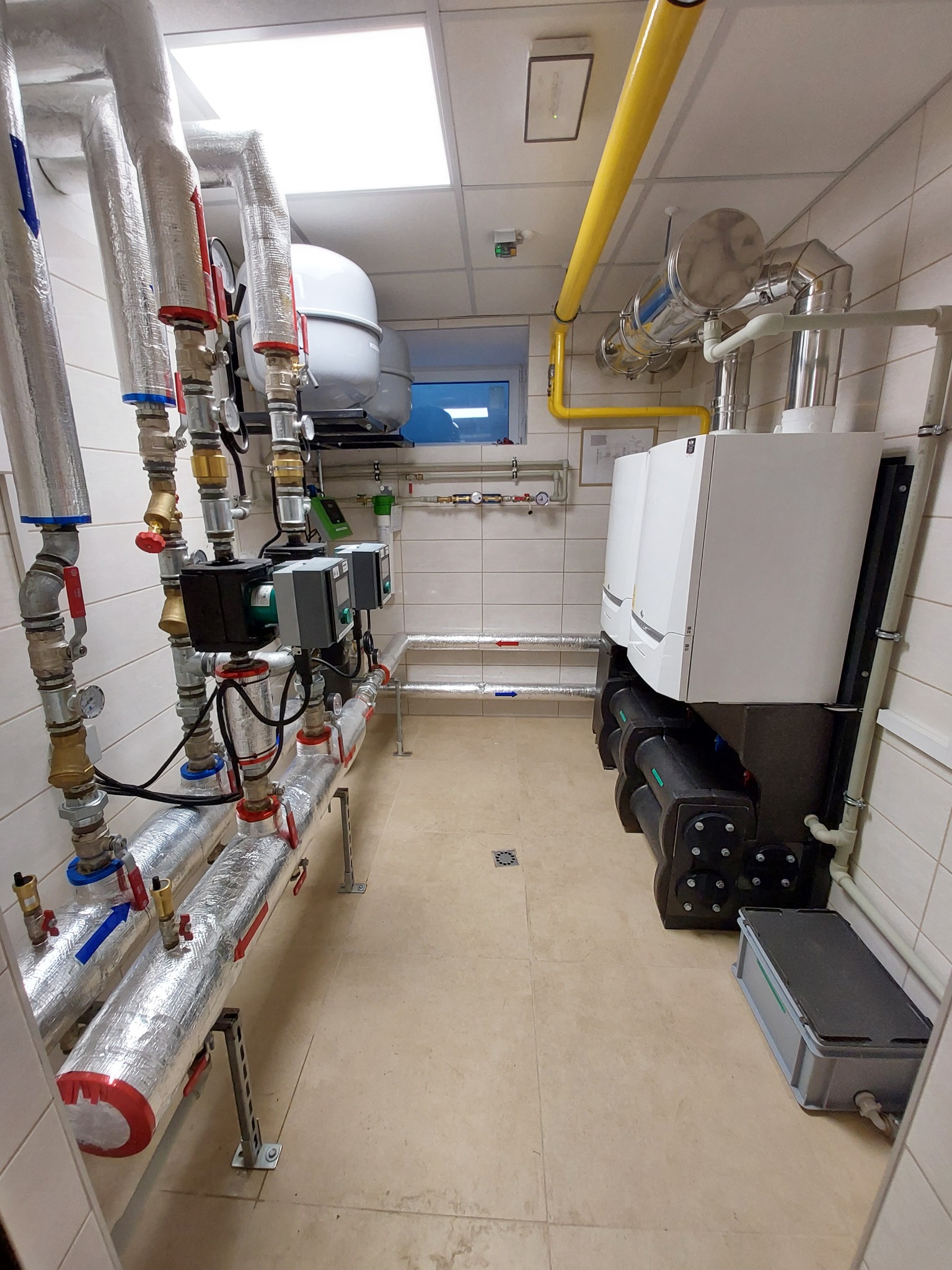 Węzeł cieplny w pomieszczeniu z dwoma białymi kotłami, orurowaniem w izolacji aluminiowej, żółtym rurociągiem gazowym pod sufitem i szarą skrzynką narzędziową na podłodze.