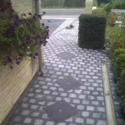 Brukowana ścieżka z kwadratowych kamieni prowadząca do domu, z donicą z fioletowymi kwiatami wiszącą na ceglanej ścianie oraz elementami kanalizacji deszczowej i roślinnością po prawej stronie.