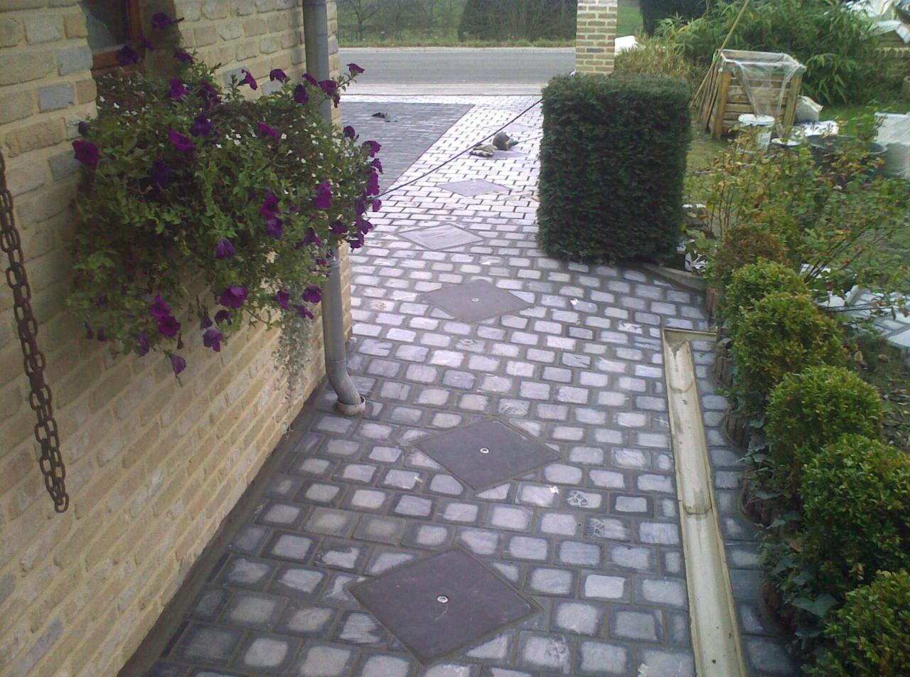 Brukowana ścieżka z kwadratowych kamieni prowadząca do domu, z donicą z fioletowymi kwiatami wiszącą na ceglanej ścianie oraz elementami kanalizacji deszczowej i roślinnością po prawej stronie.