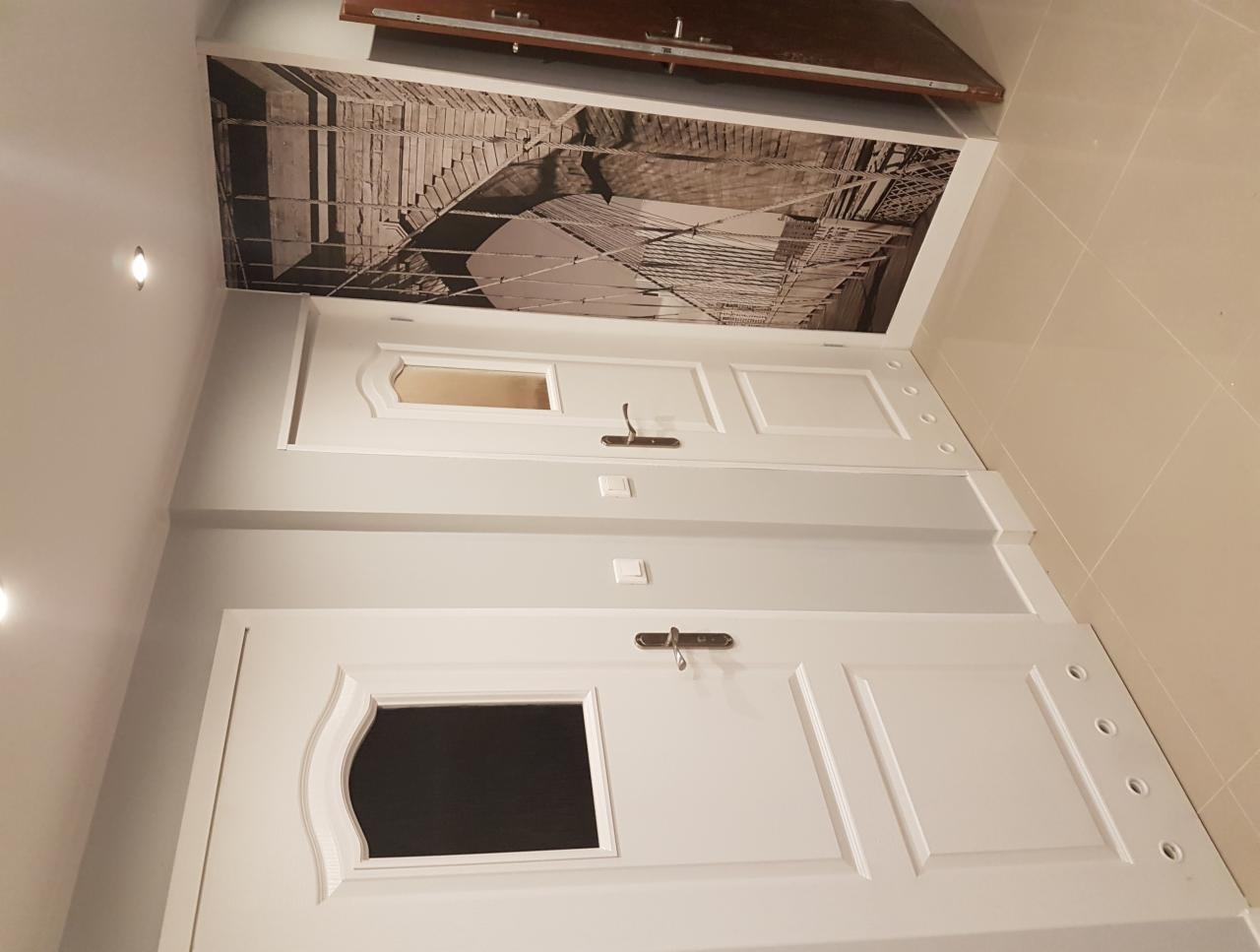 Dwa białe, identyczne drzwi wewnętrzne z przeszkleniami, zamontowane w szarej ścianie, nad nimi fototapeta z motywem mostu w stylu retro, beżowa podłoga.