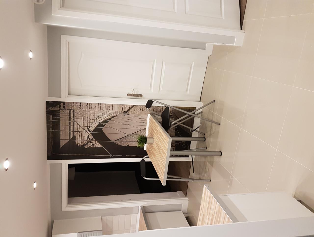 Jasne wnętrze z beżową podłogą, białymi drzwiami i dekoracyjną fototapetą z motywem mostu za stolikiem z krzesłami.