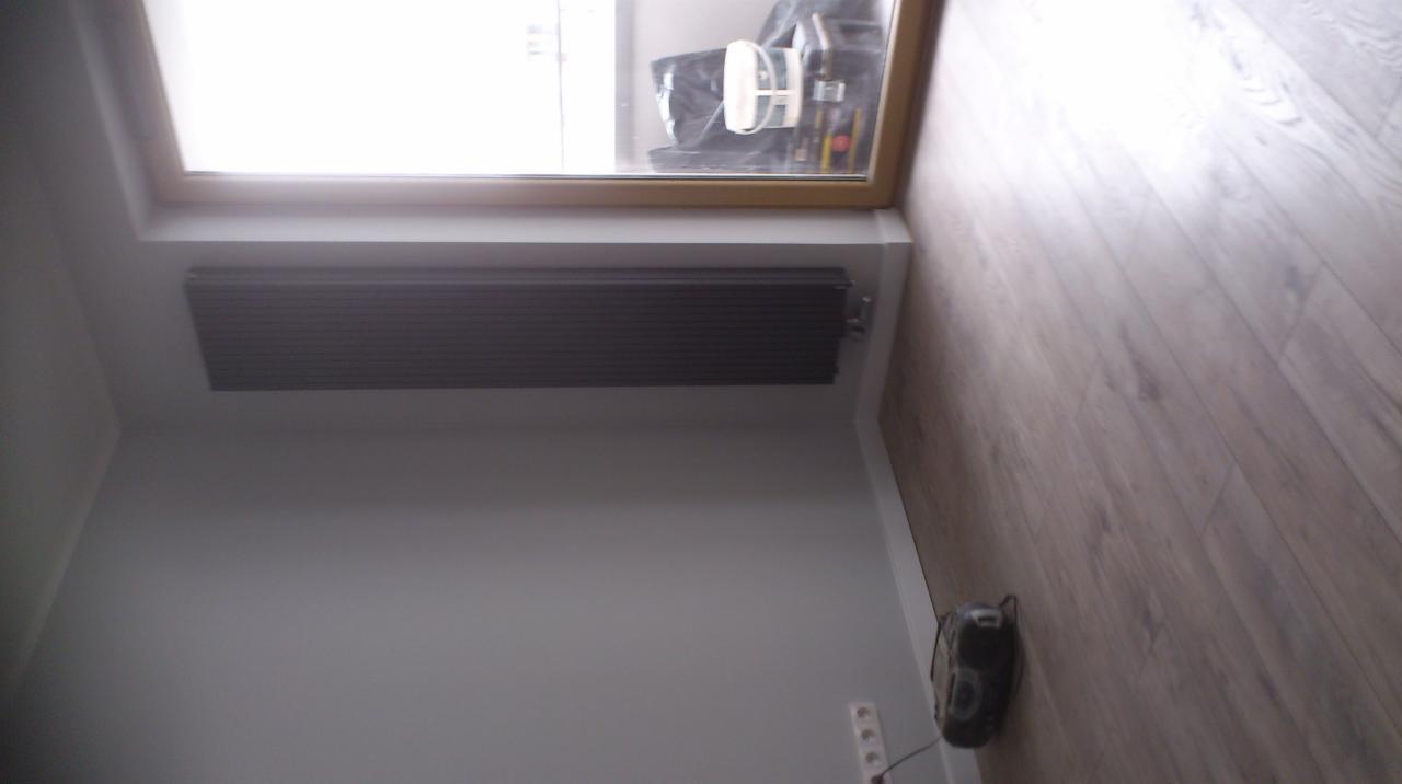 Grzejnik zamontowany pod oknem dachowym w pokoju z panelami podłogowymi, widoczny fragment ściany w kolorze szarym oraz gniazdka elektryczne.