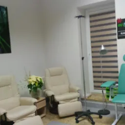 Wnętrze gabinetu kliniki ozonu i witamin: dwa jasne fotele relaksacyjne, zielony fotel zabiegowy, kroplówka, obraz z motywem leśnym na ścianie, rolety w oknie oraz szyld z nazwą kliniki.