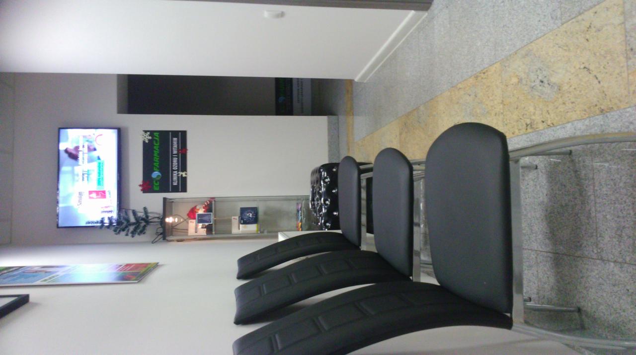 Poczekalnia w klinice ozonu i witamin, z czarnymi krzesłami, telewizorem z reklamą i półką z produktami.