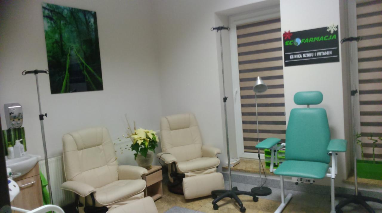 Wnętrze gabinetu kliniki ozonu i witamin: dwa jasne fotele relaksacyjne, zielony fotel zabiegowy, kroplówka, obraz z motywem leśnym na ścianie, rolety w oknie oraz szyld z nazwą kliniki.