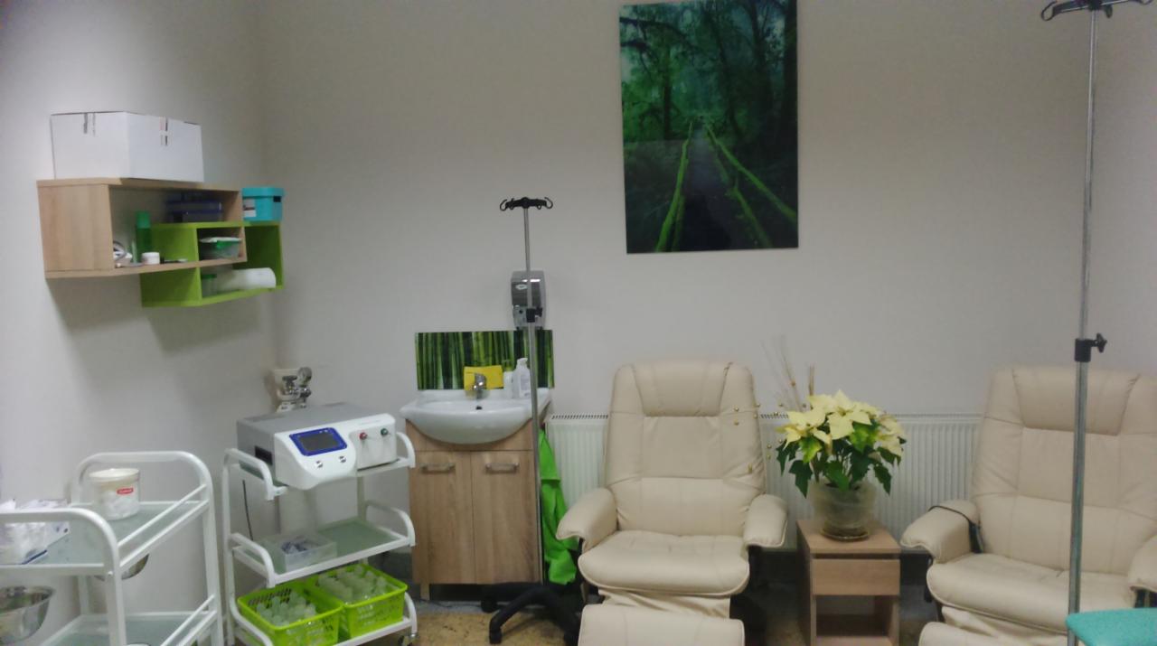 Gabinet z dwoma fotelami relaksacyjnymi, stojakiem do kroplówek, umywalką z szafką, wózkiem medycznym z wyposażeniem i obrazem leśnej ścieżki na ścianie.