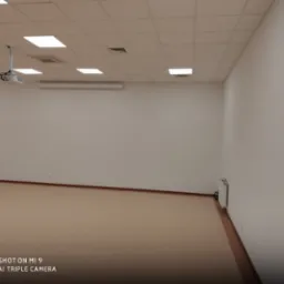 Puste, jasne pomieszczenie biurowe z beżową wykładziną, białymi ścianami i sufitem kasetonowym z wbudowanymi lampami. Na suficie widoczny projektor. Przy ścianie zamontowany grzejnik.